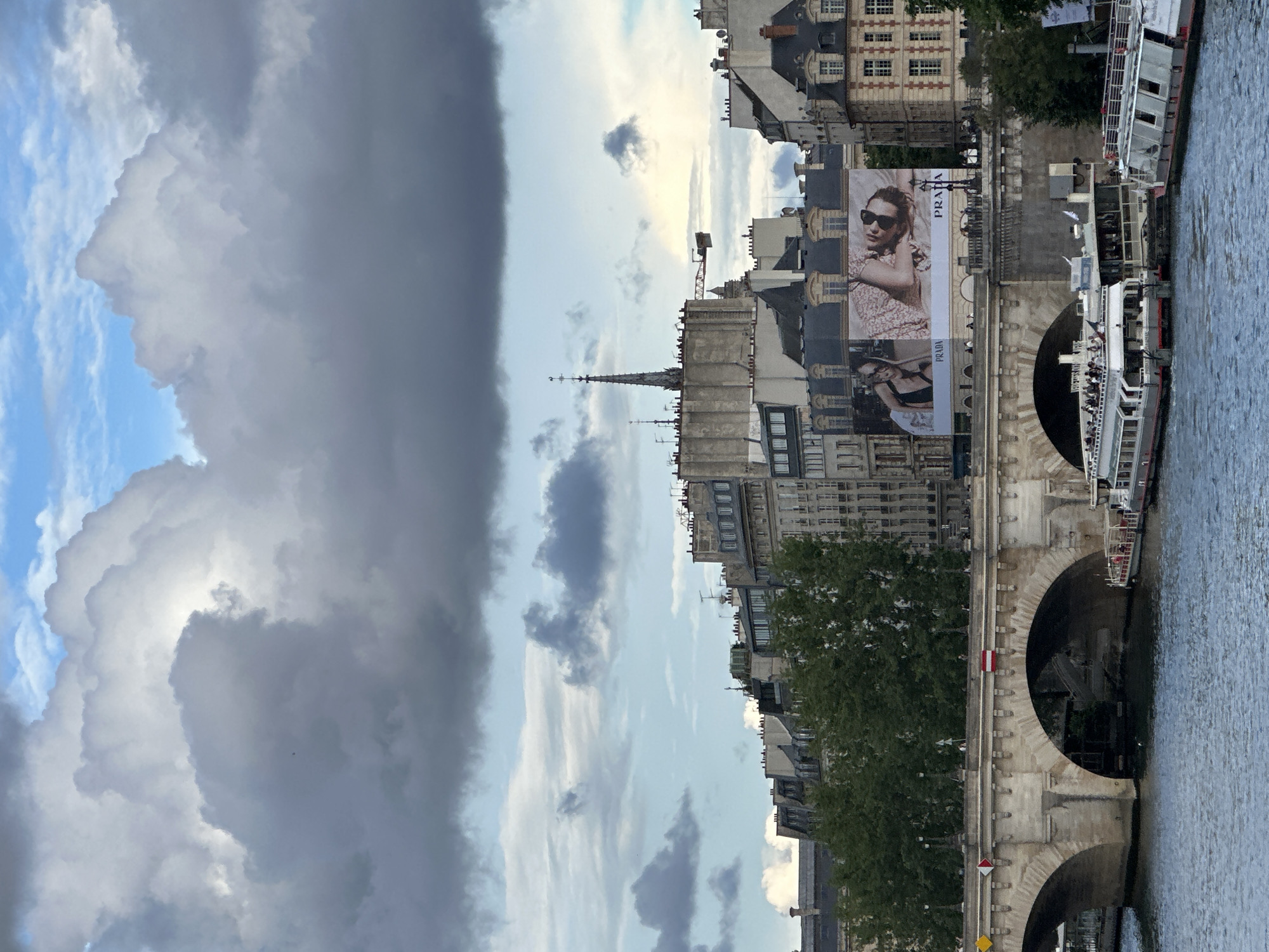 This vertical outdoor shot shows a cloudy sky above the river Seine in Paris, France. On the right, the Pont Neuf bridge, constructed of light-colored stone with visible archways, is visible. Tour boats filled with passengers are docked along the side of the bridge. Above the bridge, Parisian buildings of varying heights and architectural styles stretch into the distance. A Prada advertisement is displayed prominently on the side of a building. The river occupies the right side of the image. The sky is filled with dramatic cloud formations, ranging from light blue to dark gray.