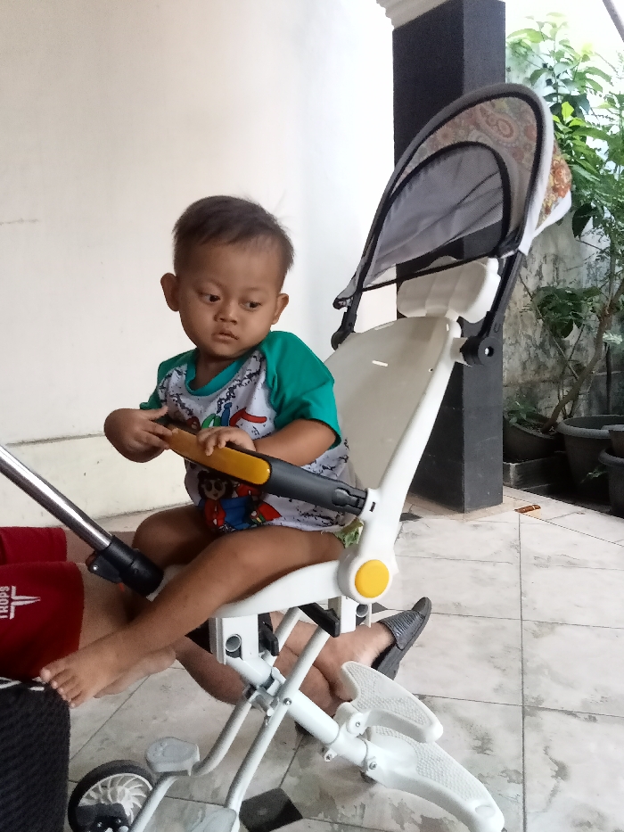 A young child, appearing to be a toddler of Asian descent, is seated in a white and black child's stroller or pushbike hybrid, featuring a gray patterned canopy and yellow frame accents. The child is barefoot, wearing a green and white long-sleeved shirt with a graphic design, and holds a yellow and black handlebar. The child looks down and to the right. The setting appears to be a tiled patio or veranda, with a plain white wall to the left and a dark gray pillar to the right. Beyond the pillar, green foliage from potted plants is visible. Part of an adult's foot, wearing a black shoe, is visible near the child's right leg. A red object with "TROPS" text is partially visible on the left. The location is an unspecified outdoor or semi-outdoor area in the city of Unknown, Unknown.