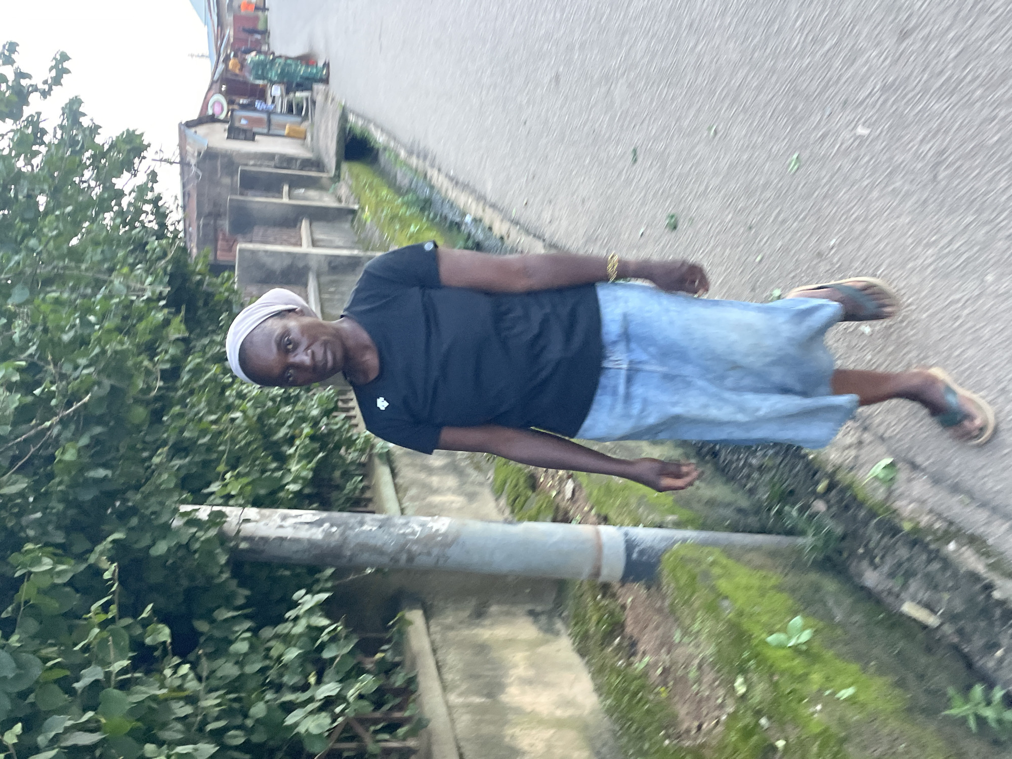 A woman wearing a black t-shirt and a light blue denim skirt walks on a paved road next to a wall covered in moss. She has a white headscarf on and is wearing flip-flops. The road is lined with trees and some low buildings. In the background, two other people are visible, one wearing a green patterned outfit. The scene appears to be in an outdoor urban setting, likely in Jos, Nigeria.