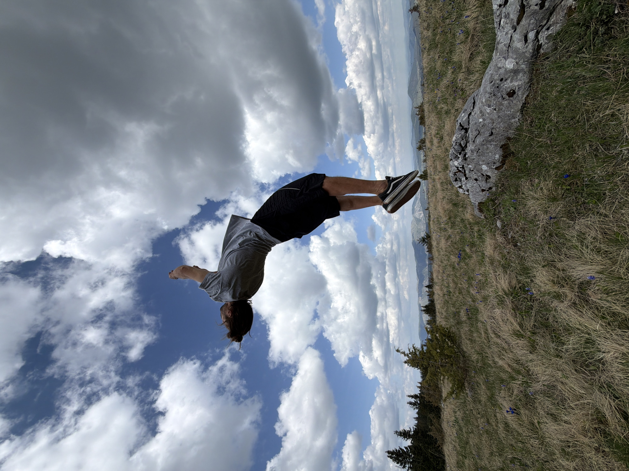 The image depicts a person in mid-air, performing a backflip. The individual is wearing a gray shirt, black shorts, and sneakers. The background includes a sky with clouds, a mountain range in the distance, and a grassy field with a large rock. The location is likely Bosača, Montenegro.
