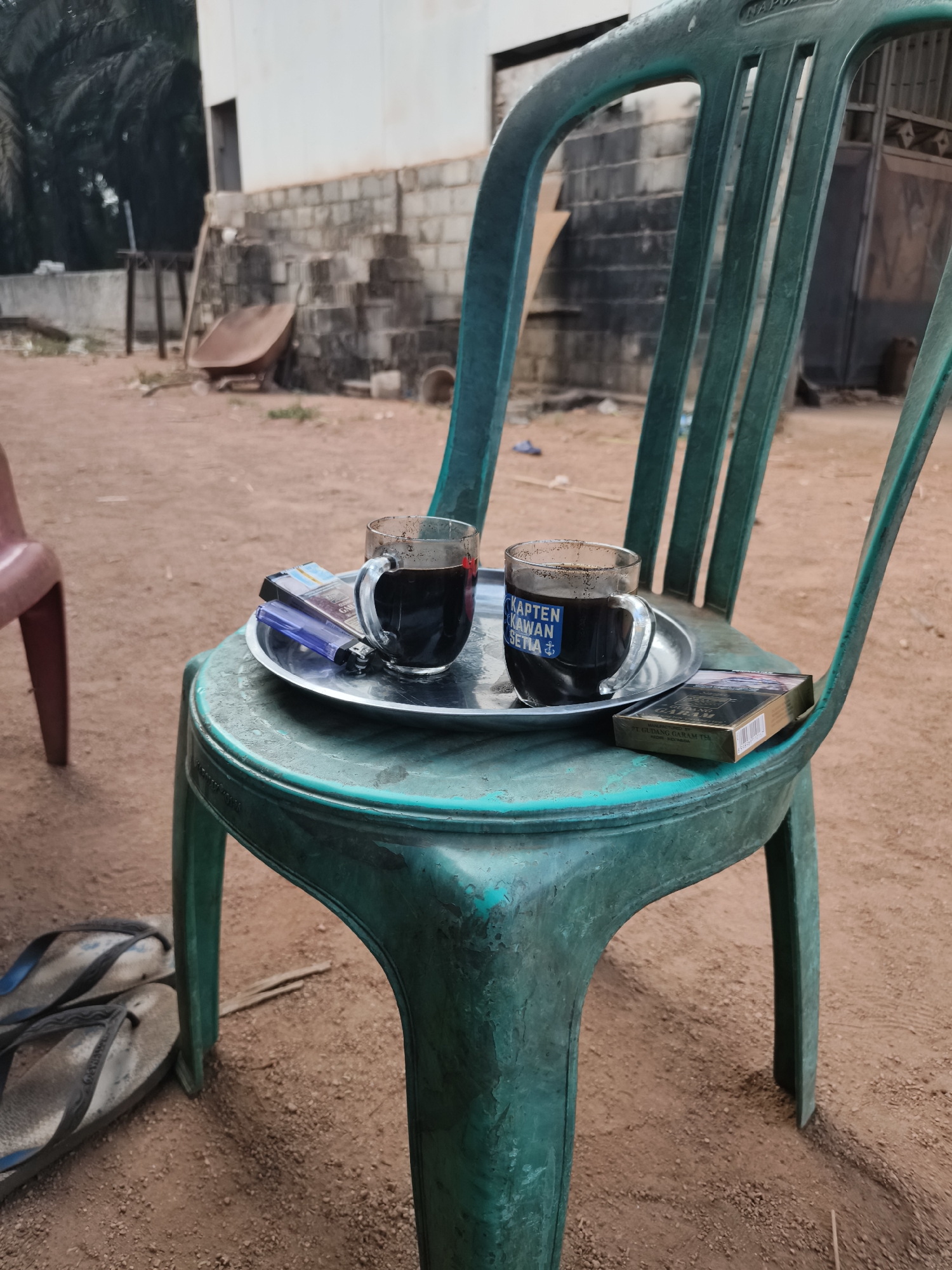 In Suka Harapan, Indonesia, two glasses of dark liquid, likely coffee, sit on a metal tray atop a green plastic chair. Beside the glasses are a lighter and a pack of Gudang Garam cigarettes. The tray and its contents are positioned on the seat of the chair, which is outdoors on a reddish-brown dirt surface. In the background, a partially constructed building with concrete blocks and stairs is visible, along with some vegetation. A pair of dirty flip-flops are on the ground to the left of the chair.