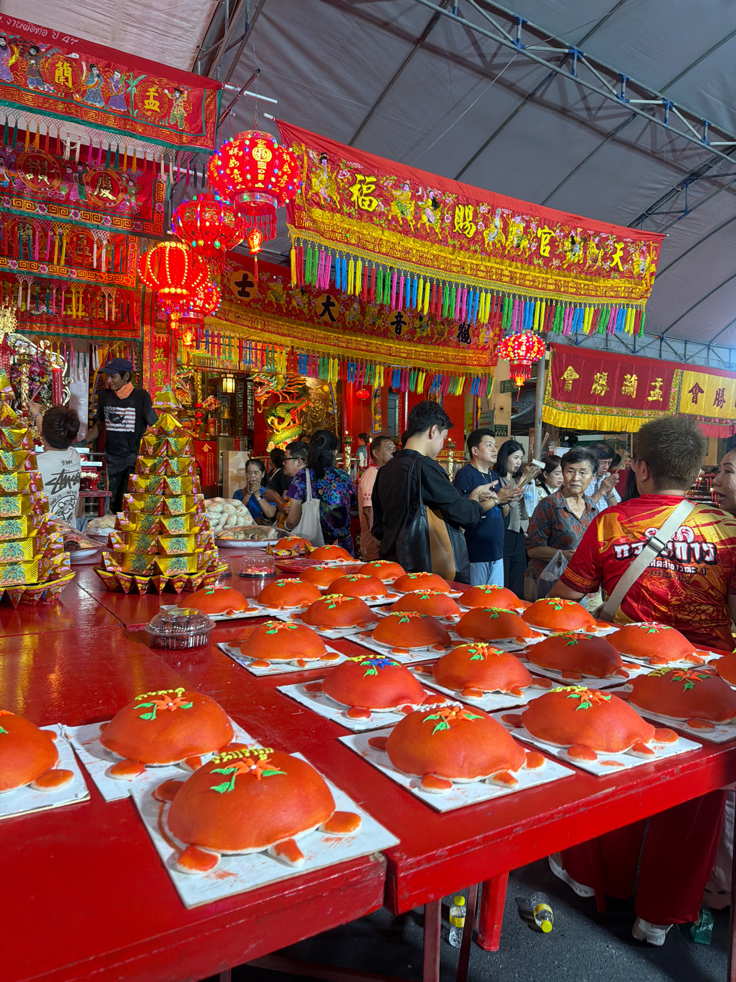 In Phuket City Municipality, Thailand, a vibrant religious or cultural event is taking place under a large tent. The scene is dominated by the color red, evident in the large red table laden with numerous orange, turtle-shaped cakes. Each cake is decorated with a small cluster of orange and green icing flowers on top, and has four small white leg-like protrusions on its sides.
Behind the table, a crowd of people are gathered. Several individuals are looking towards the front, possibly at a procession or ceremony. Some are holding their hands in a prayer-like gesture. The backdrop is adorned with ornate red and gold banners featuring Chinese characters and imagery of figures. Red lanterns, some illuminated, hang from above, adding to the festive and possibly sacred atmosphere. In the background, a temple or shrine-like structure can be glimpsed, with further decorations and possibly a dragon motif. The lighting suggests it is daytime, though the tent creates a somewhat enclosed environment. The overall impression is one of a cultural celebration with traditional offerings.