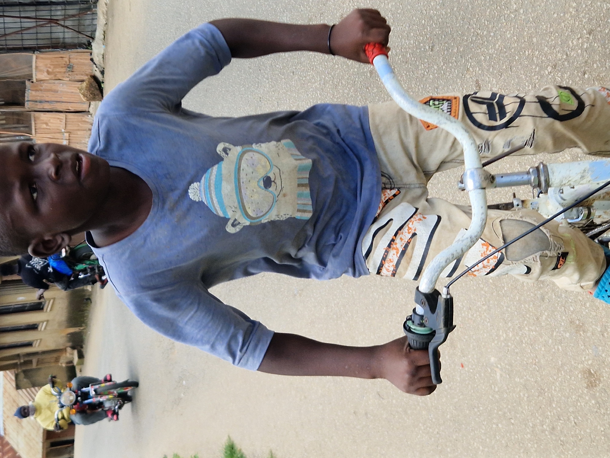 In an outdoor urban environment, a young Black male is focused on riding a bicycle. He is positioned in the foreground, facing slightly to his right, and is actively holding the handlebars of the bicycle. The boy is wearing a light blue t-shirt with a graphic of a cartoon polar bear wearing a hat and goggles, and light-colored pants with decorative patches. Behind him, another person on a motorcycle is visible, appearing to be moving away from the camera. The background features buildings with corrugated metal roofing and windows. The ground surface is paved with asphalt.