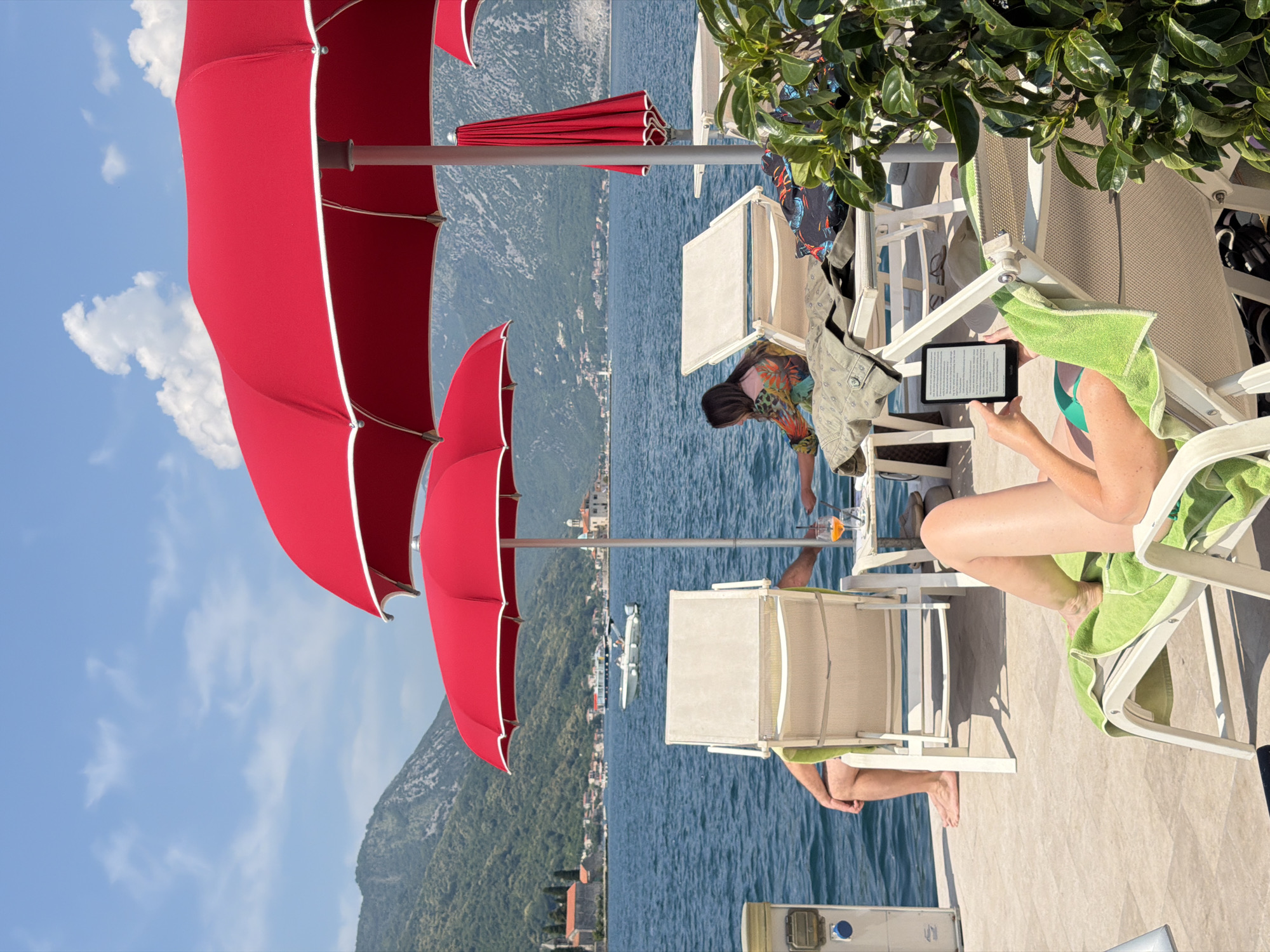 The image depicts a sunny day scene at a waterfront location, likely a pier or promenade, in Perast, Montenegro. In the foreground, a woman reclines on a green-toweled lounge chair, holding an e-reader. To her left, another lounge chair is occupied by a person's legs, with a green towel draped over it. Further back, a second lounge chair is visible, and on it, a person in a colorful shirt is reaching for a drink on a small table.
The background features the calm, blue waters of the Bay of Kotor, with a white boat sailing in the distance. Along the shoreline, traditional Montenegrin architecture, including churches and houses, is nestled against lush green hills that rise steeply. The sky is a vibrant blue with scattered white clouds.
Dominating the scene are several large, red umbrellas, providing shade over the seating area. One umbrella is fully open, while another is partially visible. A red, folded umbrella stands upright near the right edge of the frame. Lush green foliage is visible in the lower right corner, adding a touch of nature to the urban-meets-coastal setting. The overall impression is one of relaxation and enjoyment of a picturesque summer day.