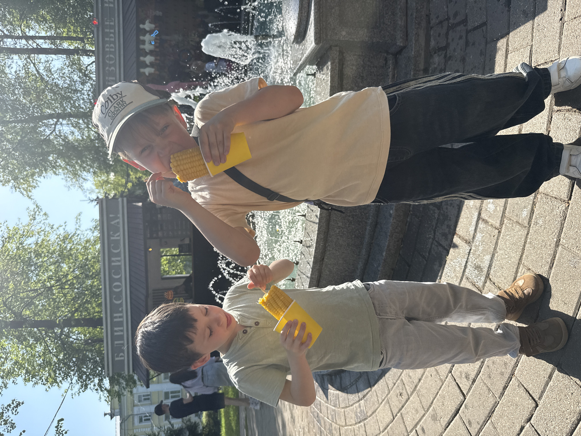 Two boys stand on a paved surface in front of a fountain in Unknown, Unknown. The older boy wears a "Colby Clark Country" cap, a light-colored t-shirt, dark pants with white-striped trim, and white sneakers; he is eating corn on the cob. The younger boy wears a light-colored shirt and light-colored pants and brown shoes and is also eating corn on the cob. Behind them is a fountain with water spraying, and a sign in Russian that translates to "Pancakes and Sausages!". Trees and buildings are visible in the background. The day appears sunny.