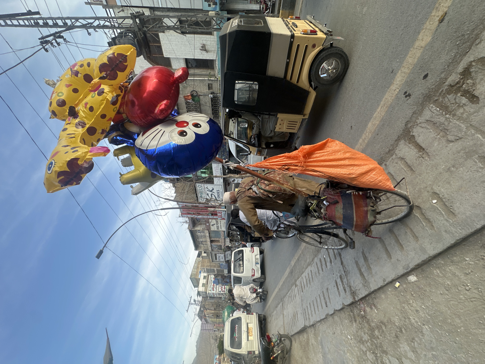 The image depicts a street scene in Quetta City Tehsil, Pakistan. A man is riding a bicycle laden with an orange bag and an assortment of colorful balloons, including yellow giraffe-shaped ones, a red round one, and a blue Doraemon character balloon. The man is wearing a light-colored shirt and cap. The background shows a street with several vehicles, including a black and beige auto-rickshaw and white vans. Buildings line the street with signs in local script. Overhead, power lines crisscross the sky. The sky is blue with some clouds. The road is paved and appears to be in average condition with some visible wear.