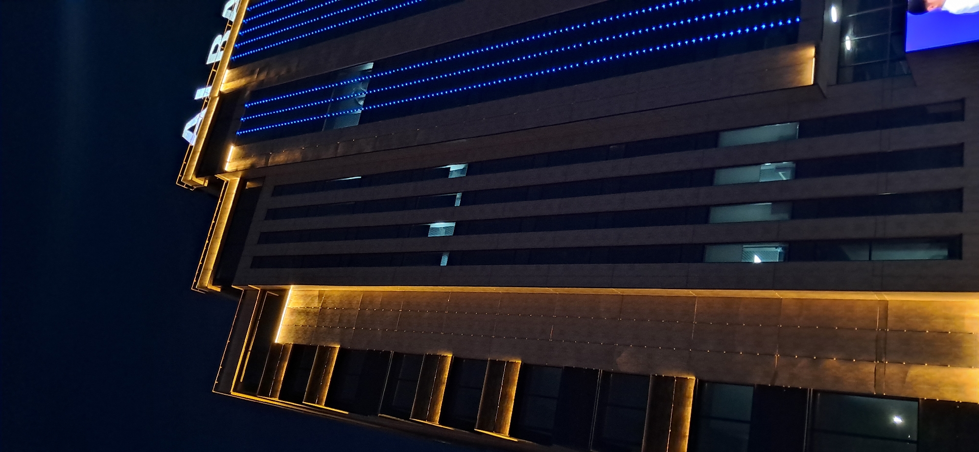 An exterior, low-angle, vertical shot captures a modern skyscraper at night, illuminated by warm golden lights accentuating its edges and vertical columns, and bright blue LED lights forming vertical stripes down its facade. The dark night sky serves as the backdrop, with the building's architecture dominating the frame. At the top of the building, the word "ALBA" is visible in illuminated white letters. Several windows on the building are lit from within, revealing glimpses of interior spaces. In the lower right corner, a large, bright blue digital screen displays a portion of a person's face. The building's construction appears to be a combination of concrete or stone panels and glass. The overall impression is one of urban sophistication and nighttime vibrancy.