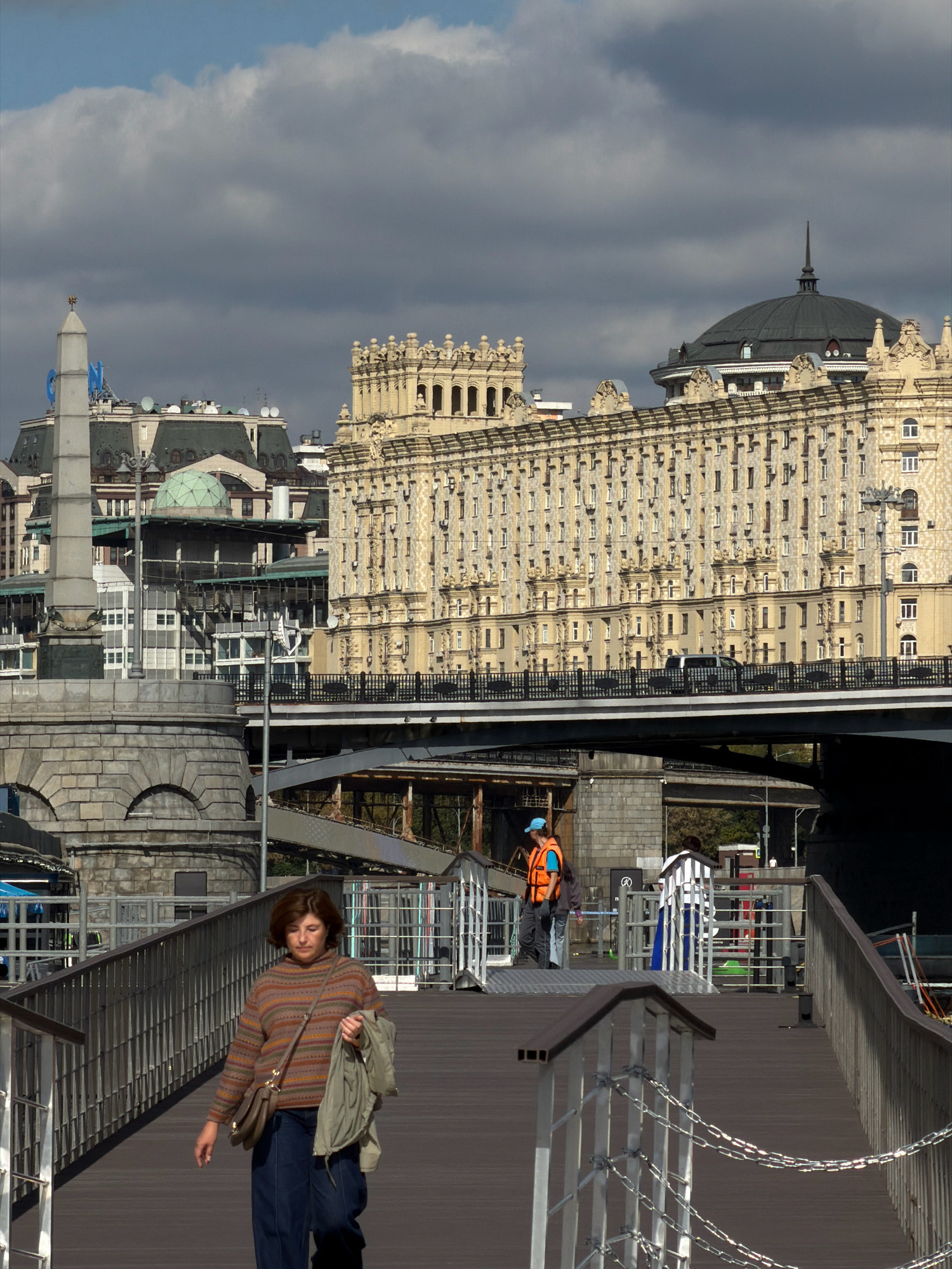 An eye-level, vertical medium shot captures a woman walking on a raised wooden walkway towards the viewer. She is dressed in a horizontally striped, multi-colored knitted sweater and dark blue pants. A brown leather purse is slung across her body, and she is carrying a folded light green jacket in her left hand. Her gaze is directed downwards. In the background, a man in an orange safety vest and a blue cap is visible on a higher level, appearing to be working. Behind him, a large, ornate building with many windows dominates the skyline, featuring a distinctive dome topped with a spire. Further in the distance, another modern building with a spire is visible. The scene is set against a backdrop of a partly cloudy sky. The walkway is bordered by a metal railing with attached chains. The overall impression is a daytime scene in an urban environment, likely a city waterfront given the presence of a bridge in the middle ground and the architectural styles suggesting Moscow, Russia.