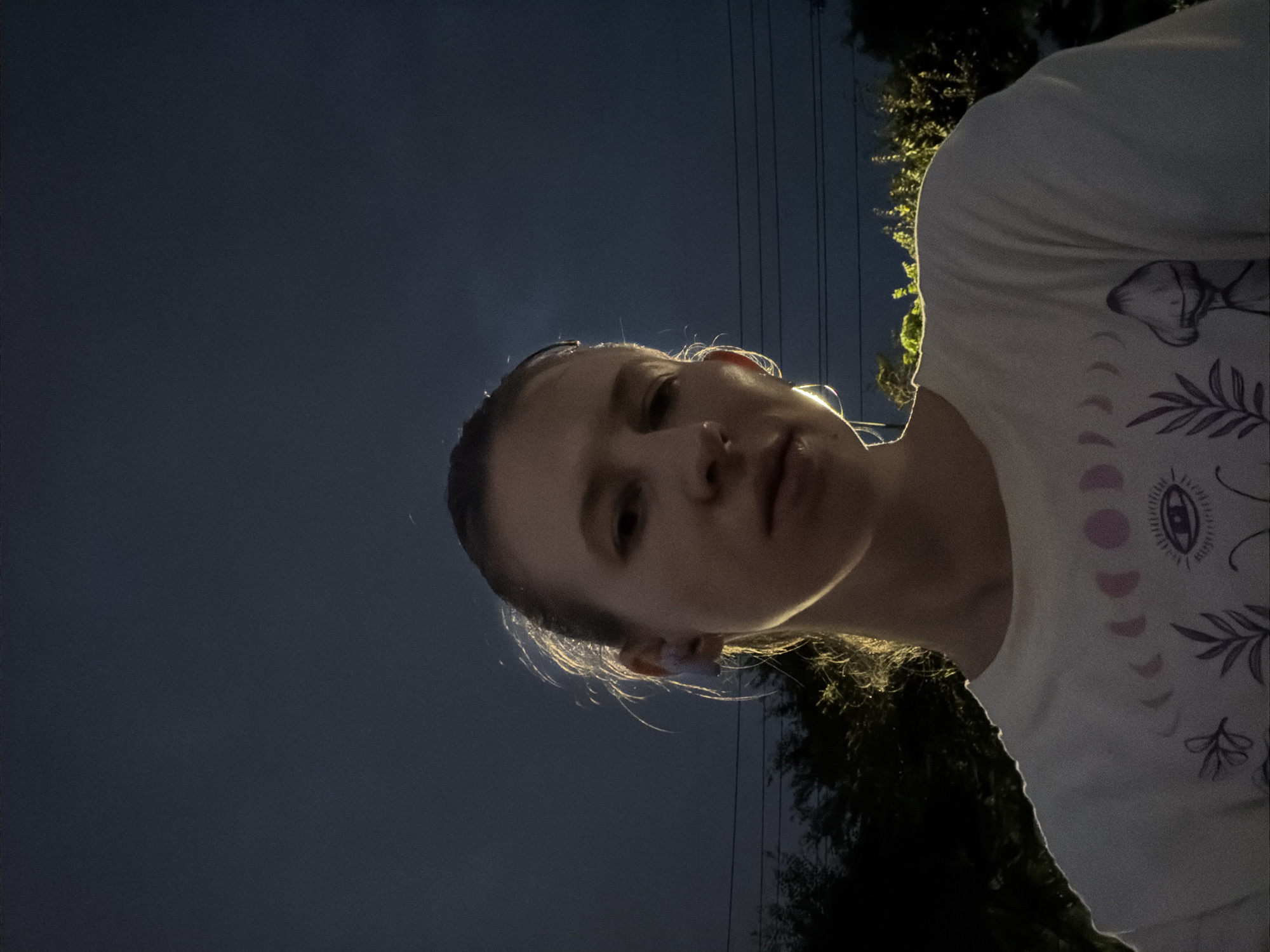 A young woman with her hair pulled back is looking forward, her face partially illuminated by a light source to her left. She is wearing a white t-shirt with a celestial and nature-themed graphic print. The background is a dark, cloudy sky with the silhouettes of trees and power lines visible at the bottom. The image appears to be a selfie taken outdoors at dusk or night. The location is not specified in the image itself, so a specific location within Dongducheon-si, South Korea, cannot be determined from the visual information provided.