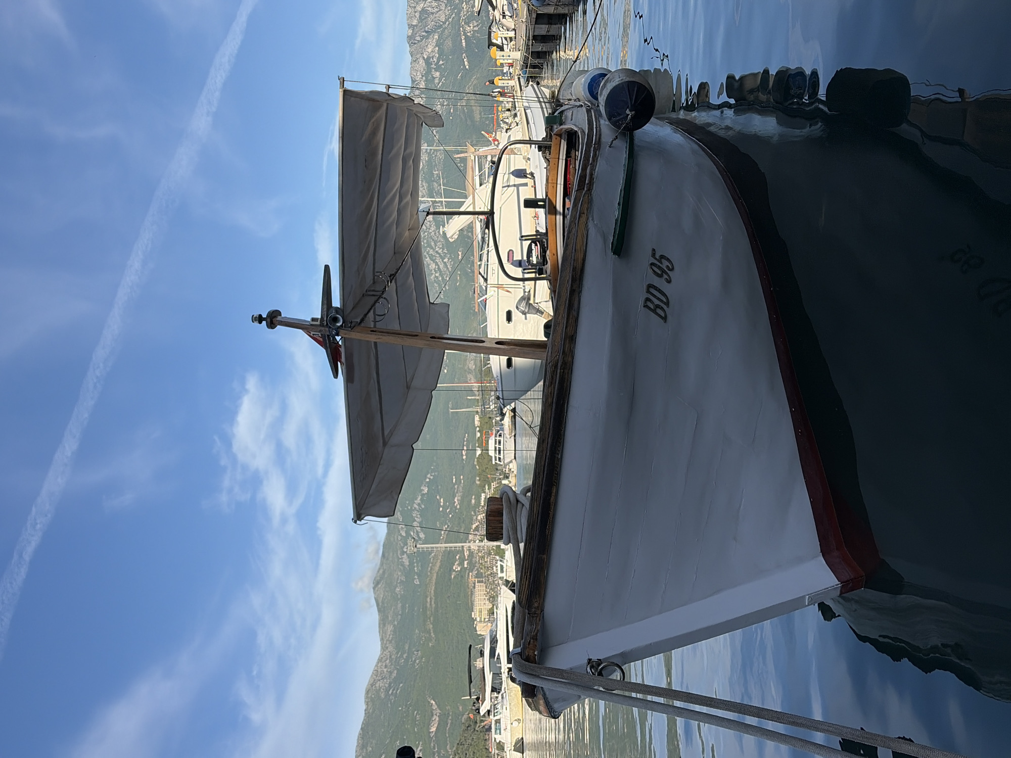 A medium shot of a boat moored at a dock in Budva, Montenegro. The boat, which has the letters "BD 95" painted on its bow, is white with a red trim. It features a small sail and various nautical fixtures. The surrounding environment includes a marina, docked boats, a mountainous backdrop, and a clear sky with visible vapor trails. The water reflects the surrounding environment.