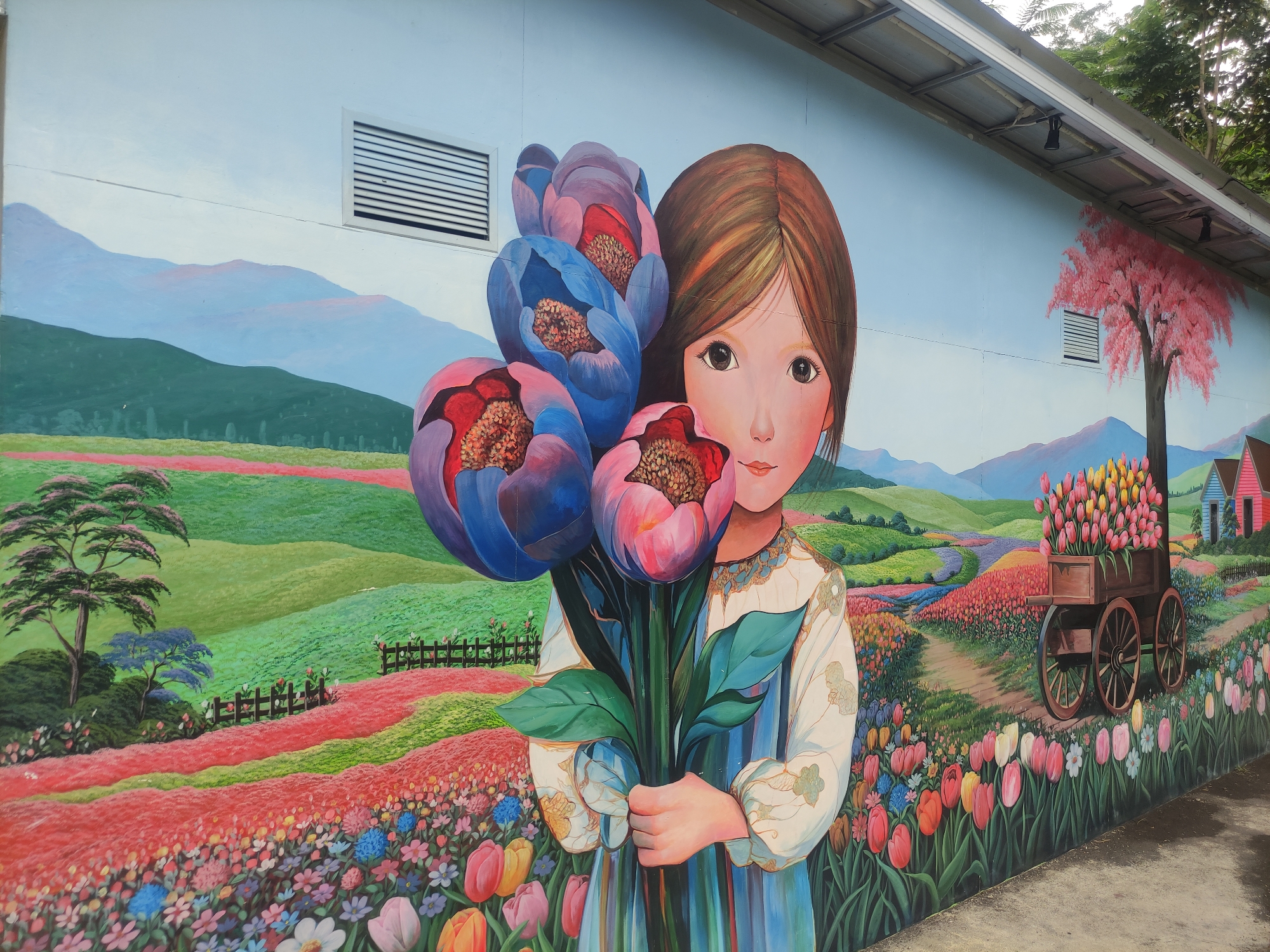 The image depicts a mural located in Babakan Madang, Indonesia. The mural is painted on a building wall. It features a young girl with brown hair and eyes holding a bouquet of oversized blue and pink flowers. Behind the girl and flowers, the background is a landscape painting depicting rolling green hills, mountains, a field of flowers, and a meandering stream. A small wooden fence appears in the background. A tree with pink blossoms and a wagon filled with tulips can be seen on the right side of the mural. The top of the building, including air vents and overhead lights, is visible above the mural.