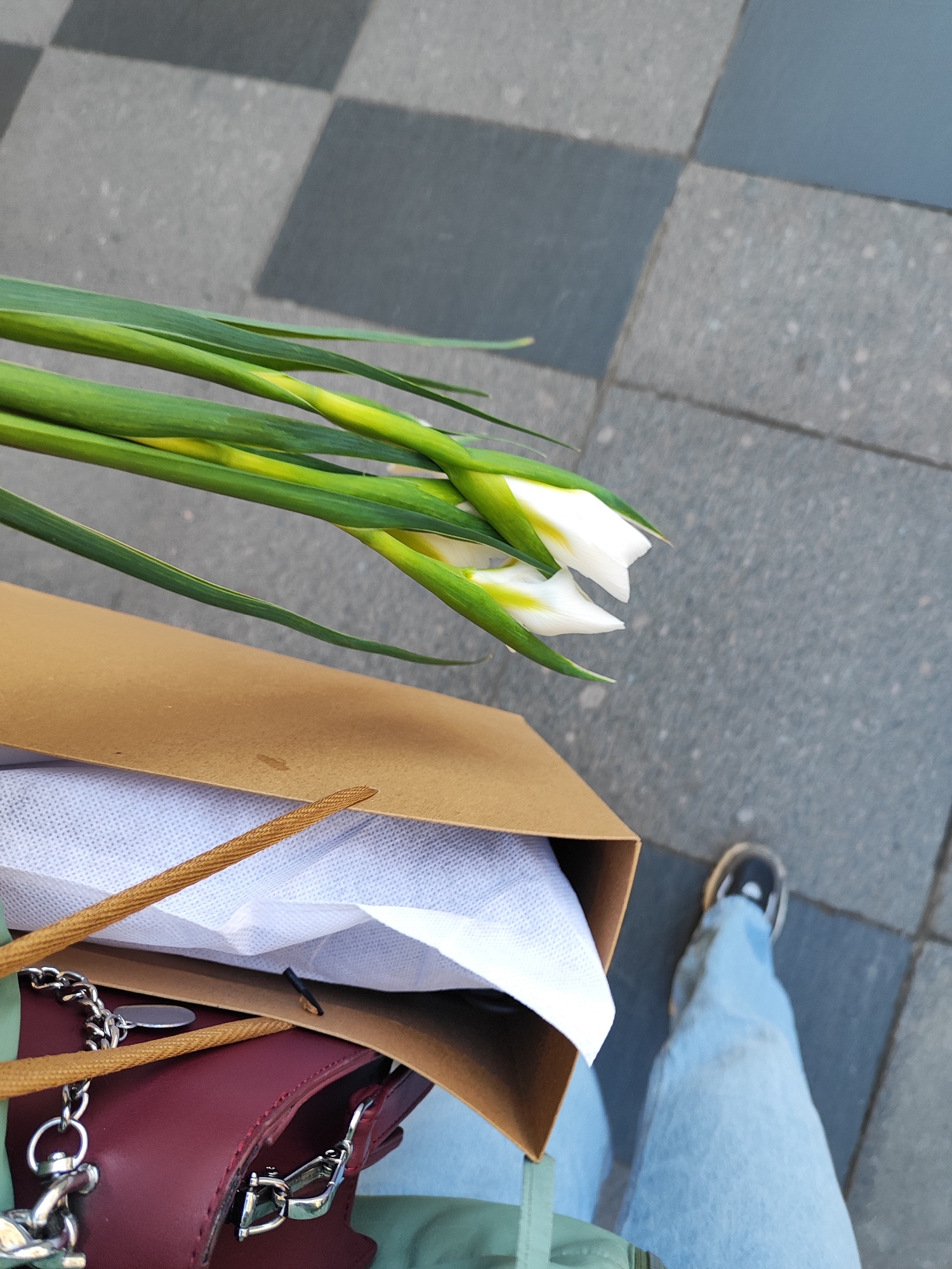 Here's a detailed description of the image:
**Scene Overview:**
The image appears to be a first-person perspective shot taken outdoors, likely on a city street with a checkered tile or paved ground surface. The angle is from above, looking down towards the subject's legs, feet, and what they are carrying.
**Subjects/Objects:**
* **Flowers:** A small bouquet of white and green flowers is visible, extending out of a brown paper shopping bag.
* **Bag:** The brown paper bag appears to contain white tissue paper.
* **Handbag:** A burgundy handbag with silver hardware and a chain is visible beneath the shopping bag.
* **Clothing:** The person is wearing light blue jeans and black and white sneakers. A glimpse of a light green garment is visible on the torso.
* **Ground:** The ground surface is comprised of grey tiles in a checkerboard pattern.
**Actions/Interactions:**
* The person is holding the bag with the flowers and the burgundy handbag.
* Based on the angle, it is probable that the person is walking.
**Environment:**
* **Location:** The location seems to be an urban environment, given the pavement style.
* **Lighting:** The lighting appears to be natural daylight, although there is no strong indication of time of day.
The image conveys a feeling of casualness and an everyday moment, possibly a person returning home after shopping or receiving flowers.