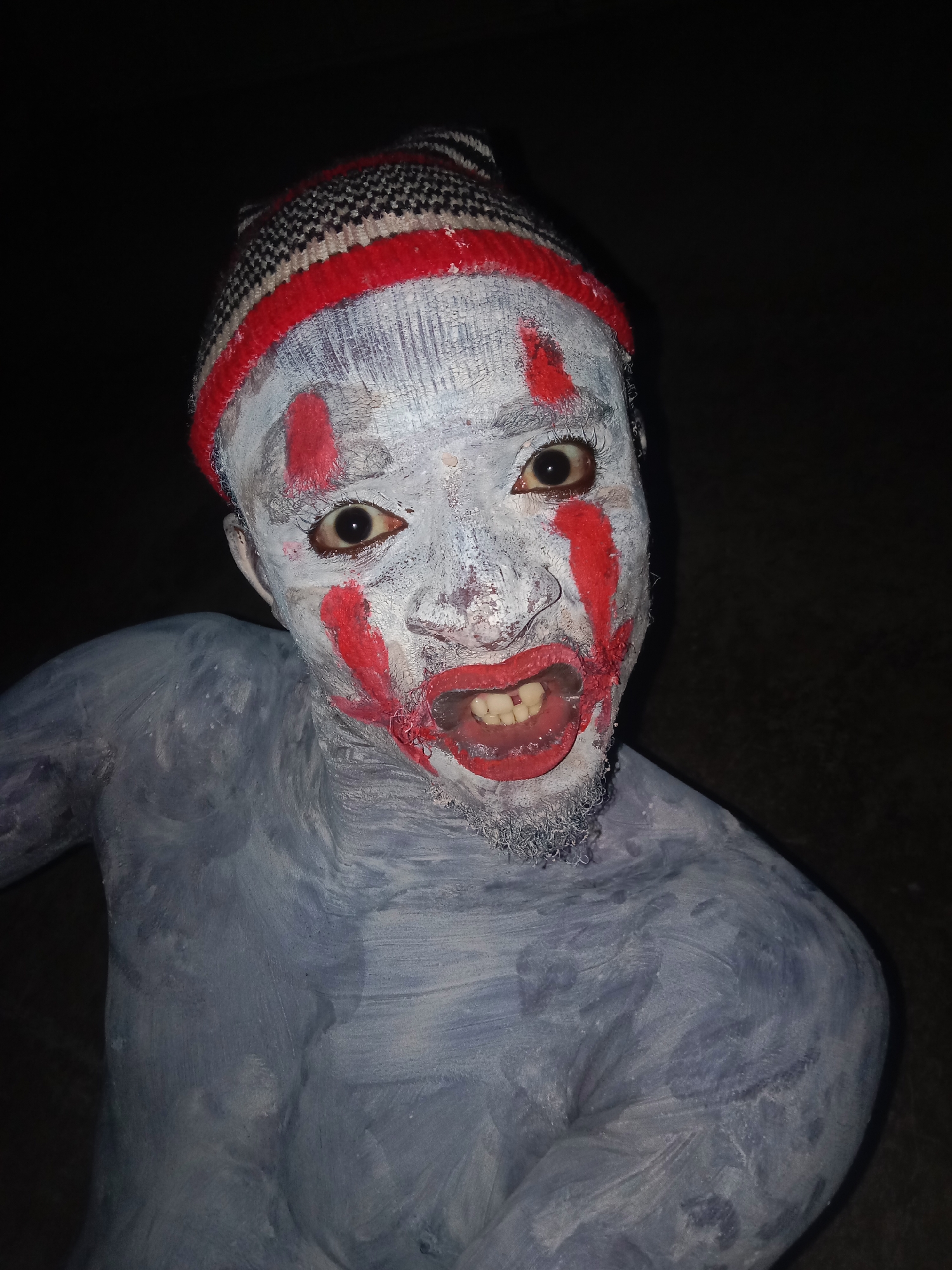 A person with their face and body painted white and red, wearing a striped beanie, is looking directly at the camera. The background is dark, making it difficult to discern specific details about the location. The city and location are unknown.
