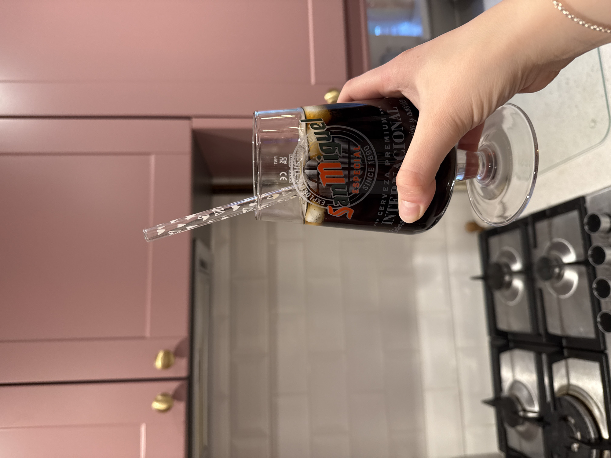 A fair-skinned hand is holding a glass of San Miguel beer. The glass has a clear straw with white heart patterns in it. The background includes light pink cabinets with gold knobs and a white tiled wall, and a stainless steel stovetop. The image appears to be taken indoors in a kitchen. It is not possible to determine the location in the city of East Suffolk, United Kingdom.