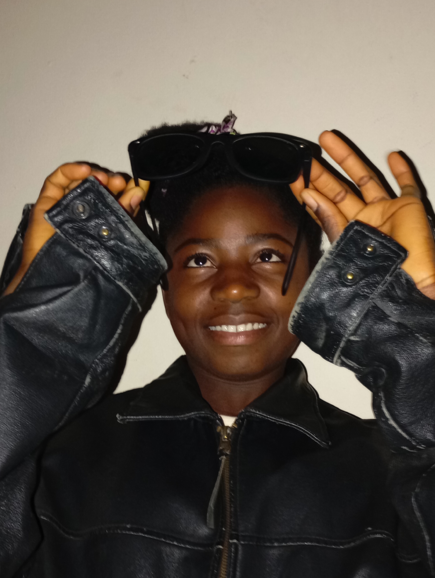 A person with dark skin and short black hair is holding sunglasses on top of their head. They are wearing a black leather jacket. The background is a plain white wall. The location is given as Jos, Nigeria.