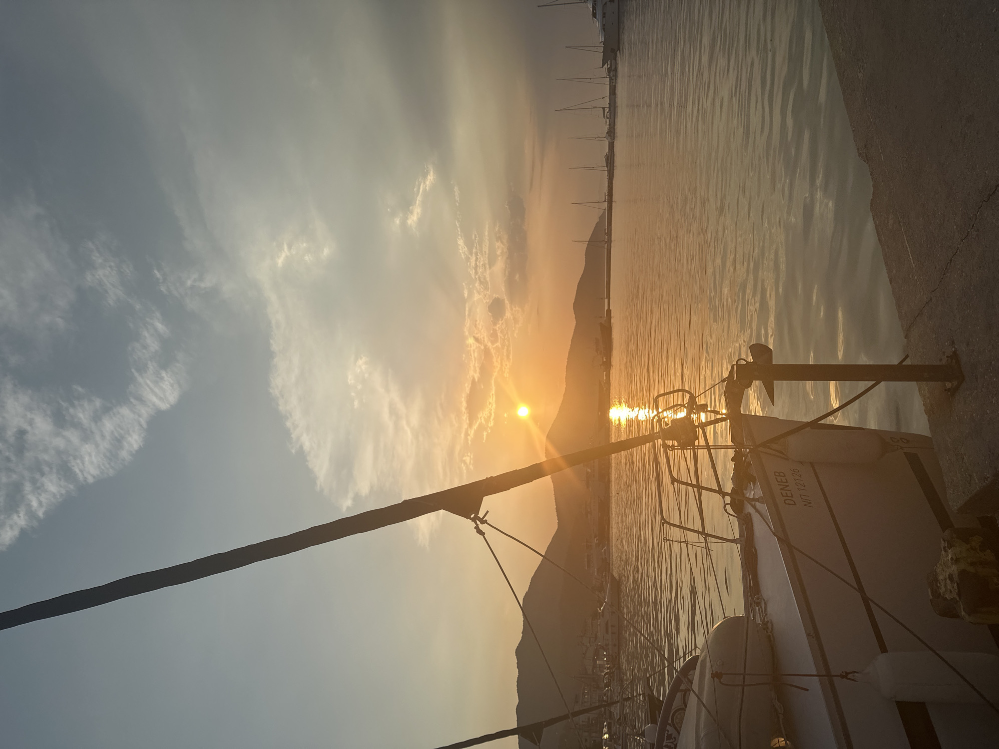 The image shows a sunset over the harbor in Thassos, Greece. The sun is low in the sky, casting a warm, golden light over the water and the surrounding hills. Several sailboats are docked in the harbor, their masts silhouetted against the colorful sky. In the foreground, the bow of a sailboat is visible, with the name "DENEB" and the registration number "NII 12126" painted on its hull. The water is calm, reflecting the colors of the sunset. The scene is peaceful and serene, capturing the beauty of a Mediterranean evening.
