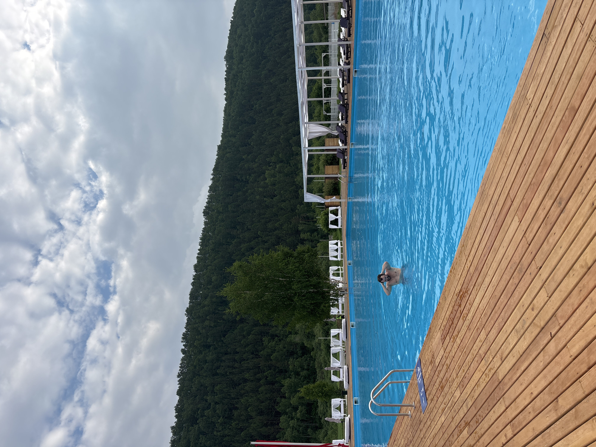 A medium shot captures a person standing in the middle of a bright blue swimming pool at a resort in the city of Дрокино, Russia. The person, whose head and shoulders are visible above the water, has their hands near their head. The pool is bordered by a wooden deck in the foreground, with a silver ladder visible on the left. In the background, a line of white cabanas with white curtains stands along the pool's edge, some of which have lounge chairs in front of them. Beyond the cabanas, a dense green forest covers a large hill, and the sky above is filled with white, fluffy clouds. The overall scene suggests a leisurely day at an outdoor swimming facility.