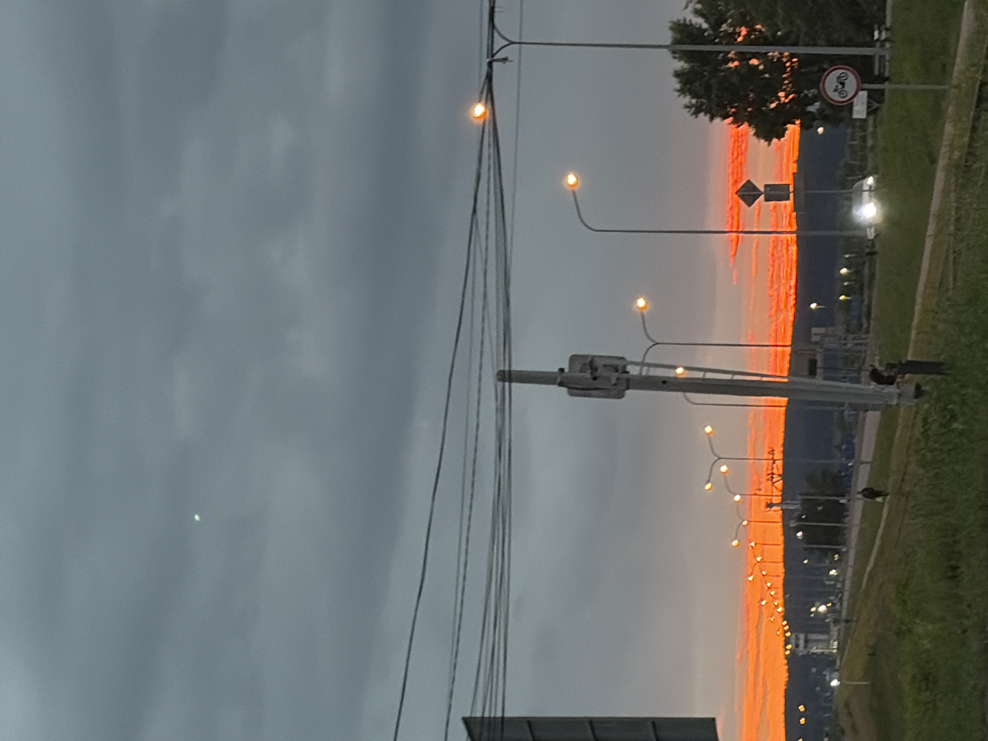 This image depicts a roadside scene at dusk, with a vibrant orange and red sunset dominating the horizon. The sky is partly cloudy, with darker, gray clouds in the upper portion and a bright, fiery glow where the sun is setting. A row of streetlights line the road, some of which are illuminated. In the foreground, there is a grassy embankment and a paved pathway.
Along the road, several objects are visible:
- A tall signpost with multiple signs, including a diamond-shaped warning sign and a circular sign with a motorcycle symbol, indicating a prohibition.
- Electrical wires stretch across the upper part of the frame, supported by poles.
- A few vehicles are present on the road, with headlights illuminated.
- Several indistinct figures of people can be seen walking or standing along the road and pathway.
- A small, bright green light or object is visible in the upper left quadrant of the sky.
The location is identified as Chaikovsky, Russia, specifically suggesting a view along a road with street lighting and a sunset, possibly on the outskirts of the city given the presence of a natural horizon and the road infrastructure. The overall atmosphere is tranquil, with the dramatic sunset providing a striking visual element.