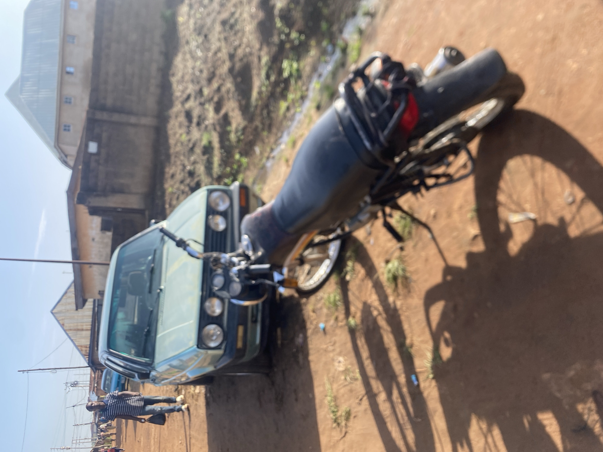 In Bukuru, Nigeria, a man walks on a dirt road in front of a light green Volkswagen Golf. A black motorcycle is parked parallel to the car. In the background, there are buildings and more people walking on the street. The sky is clear and blue, and the sun is shining.