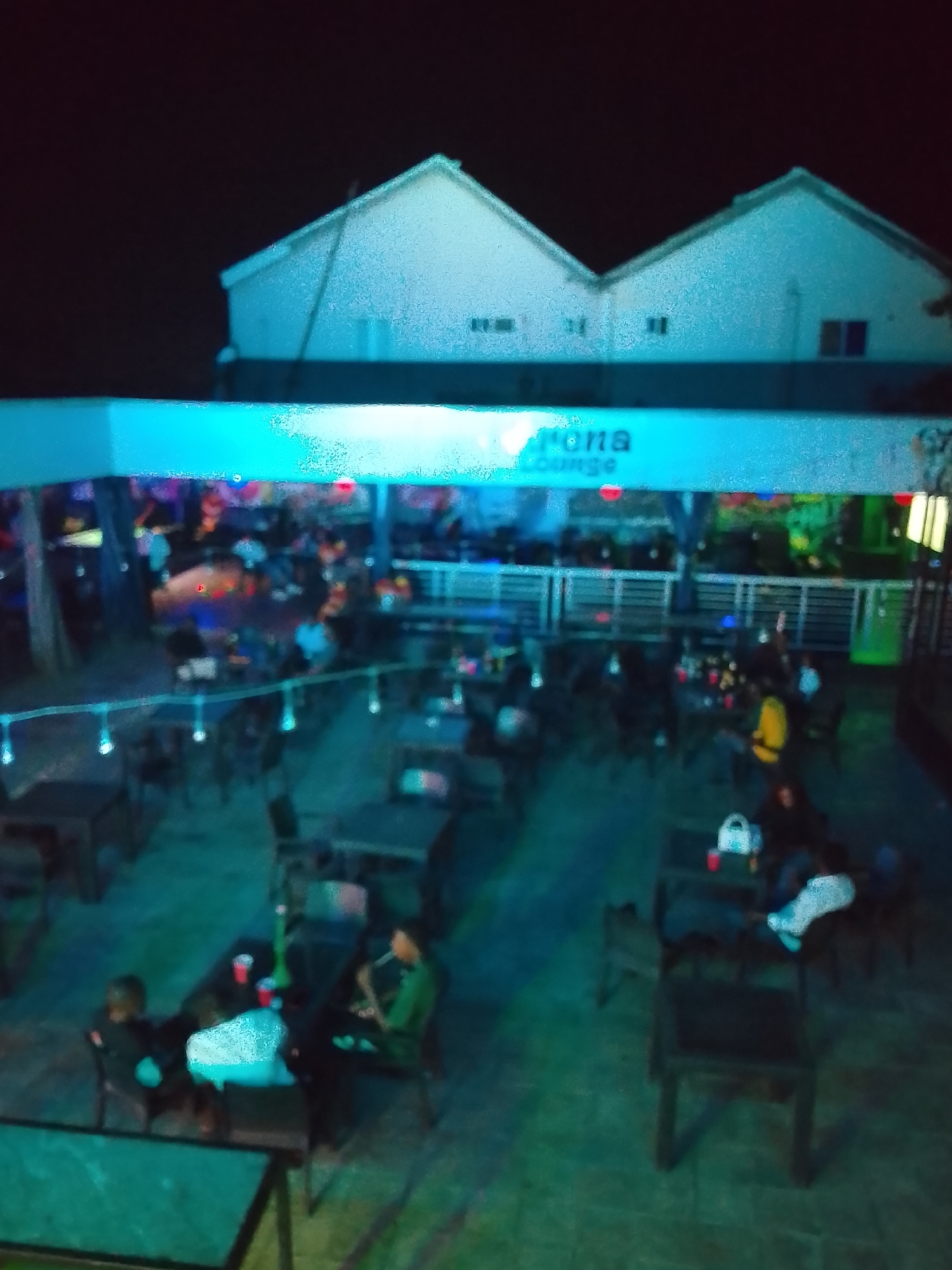 The image shows the exterior of the Corona Lounge in Narayi, Nigeria at night. The lounge has a covered outdoor seating area with numerous tables and chairs, occupied by patrons. String lights are hung above the seating area. The lounge name, "Corona Lounge", is visible on the roof of the seating area. Behind the lounge, the silhouettes of residential buildings are seen against the dark sky. The overall lighting is low, and the image appears slightly blurry.