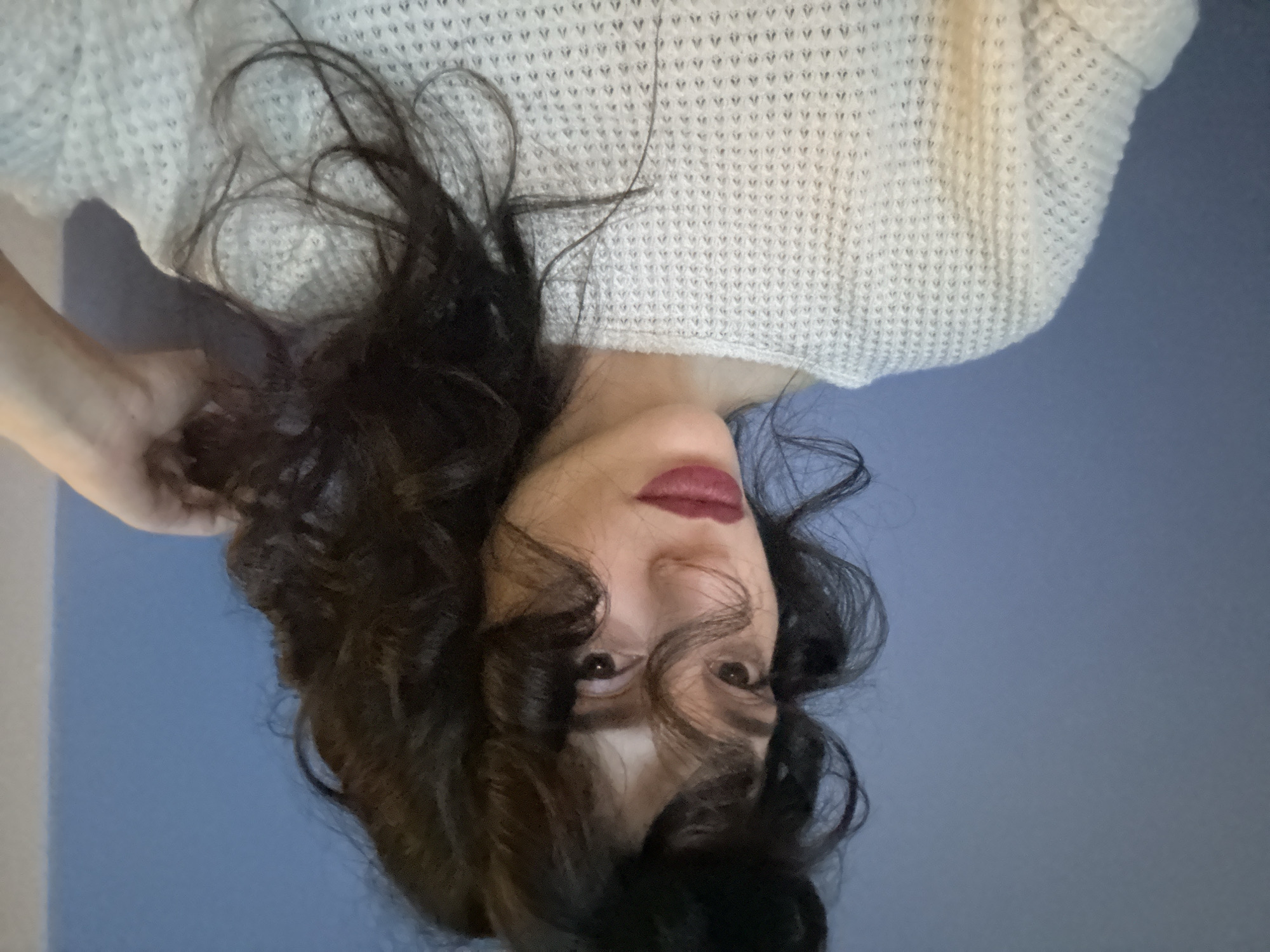 This media file features a close-up, upside-down shot of a person's face and upper body, with a focus on their features and clothing. The subject has dark, wavy hair that frames their face and is being touched by their left hand. They are wearing a white, textured sweater, possibly a knit. The background is a solid, muted blue wall, with a hint of a lighter surface on the far left, possibly a desk or furniture.
The lighting appears to be artificial and soft, creating subtle shadows on the face and neck. The subject's eyes are open and looking towards the camera, with a neutral or slightly contemplative expression. Their lips are colored with a dark lipstick. The overall mood is intimate and personal due to the close-up framing and the way the hair is styled.
There is no visible text in the image. The setting suggests an indoor environment, likely a home or a studio, given the plain background and casual attire. The time of day and weather are not discernible from the image. The location context of Sofia, Bulgaria, does not provide any specific visual cues within this particular frame.
