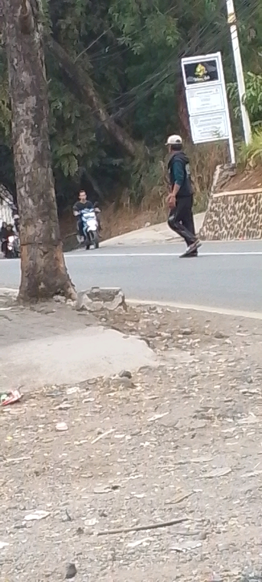 In Cilaku, Indonesia, a man wearing a cap and a dark vest over a teal shirt and dark pants is walking across a paved road. A motorcycle with a rider in a dark shirt is approaching him from the left. In the background, a sign with a golden emblem and text is mounted on a pole. The road is bordered by a stone wall on the right and trees and foliage on the left. The foreground is rough terrain with scattered debris.