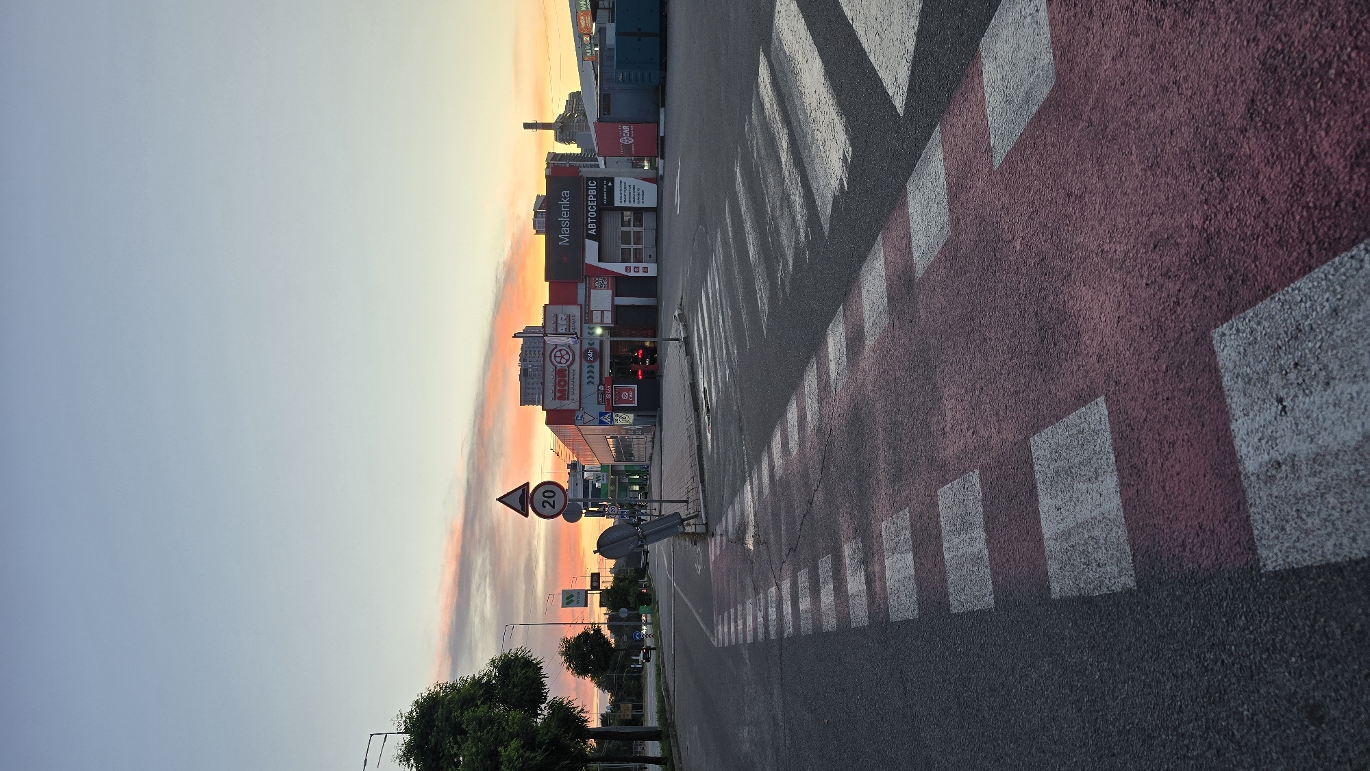 The image depicts a pedestrian crossing on a street in Kyiv, Ukraine, during a sunrise or sunset. The sky displays hues of orange, pink, and blue, indicating transitional light conditions. On the right side of the street, there is a commercial building complex with signage including "MASLENKA" and "АВТОСЕРВІС," suggesting it houses a service station or car-related business. Traffic signs, including a speed limit sign indicating "20," are visible. A few cars are present on the road, and trees line the left side of the street. The pedestrian crossing itself is marked with white and red painted stripes on the asphalt. The scene appears to be an urban environment at either the beginning or end of the day.