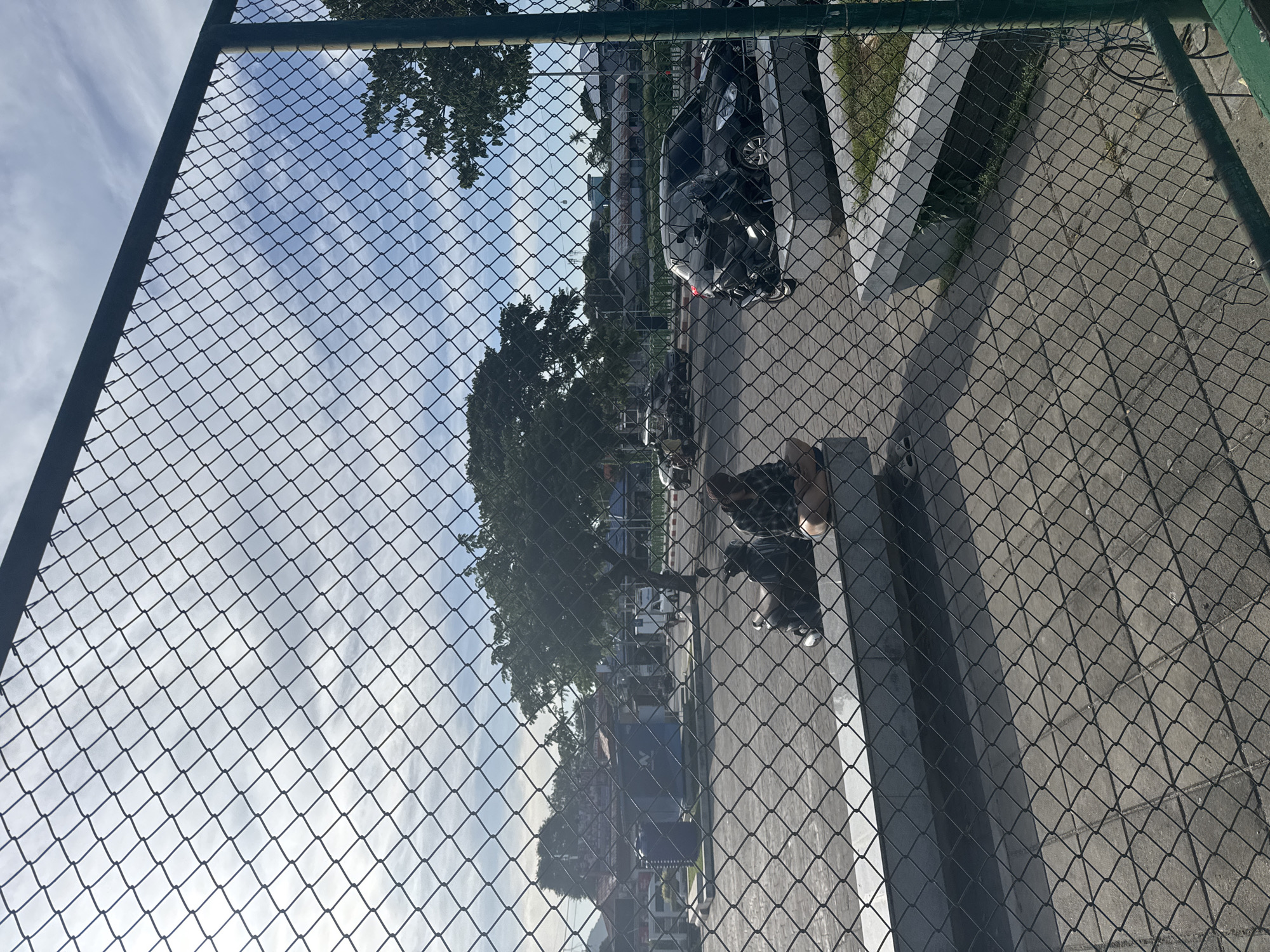 In Ko Samui District, Thailand, a chain-link fence obscures a view of a paved outdoor area. Two individuals, one wearing a checkered shirt and the other a dark top, sit on a raised concrete bench. Next to them is a parked motorcycle. In the background, several vehicles are visible, including a black SUV, another black car, and a motorcycle. The scene is set against a backdrop of trees and buildings under a partly cloudy sky.