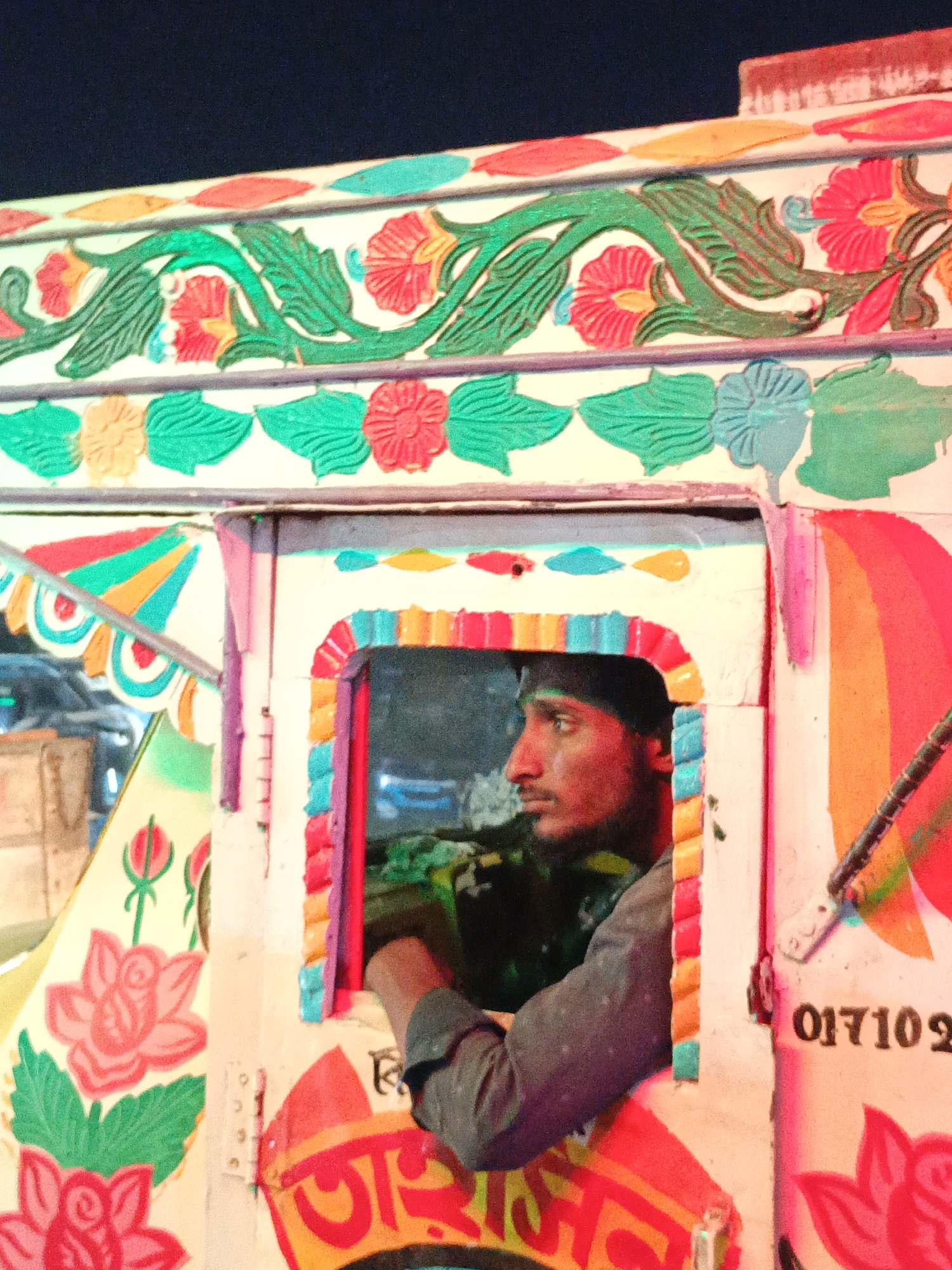 A medium, eye-level shot shows a man in profile looking out from the window of a brightly colored vehicle. The man has dark skin and wears a dark hat and a gray shirt. He has his arm resting on the window frame. The window frame is decorated with bright pink, blue, yellow, and orange patterns. The vehicle itself is white with floral designs in red, green, and teal. There is a row of stylized flowers and leaves along the top of the vehicle. A rainbow design can be seen to the right of the window. A number is partially visible on the right side of the vehicle: "017102". The interior of the vehicle is dark. The background is dark and blurry, suggesting a nighttime scene. Some elements suggest this might be a painted rickshaw. The location is unknown.