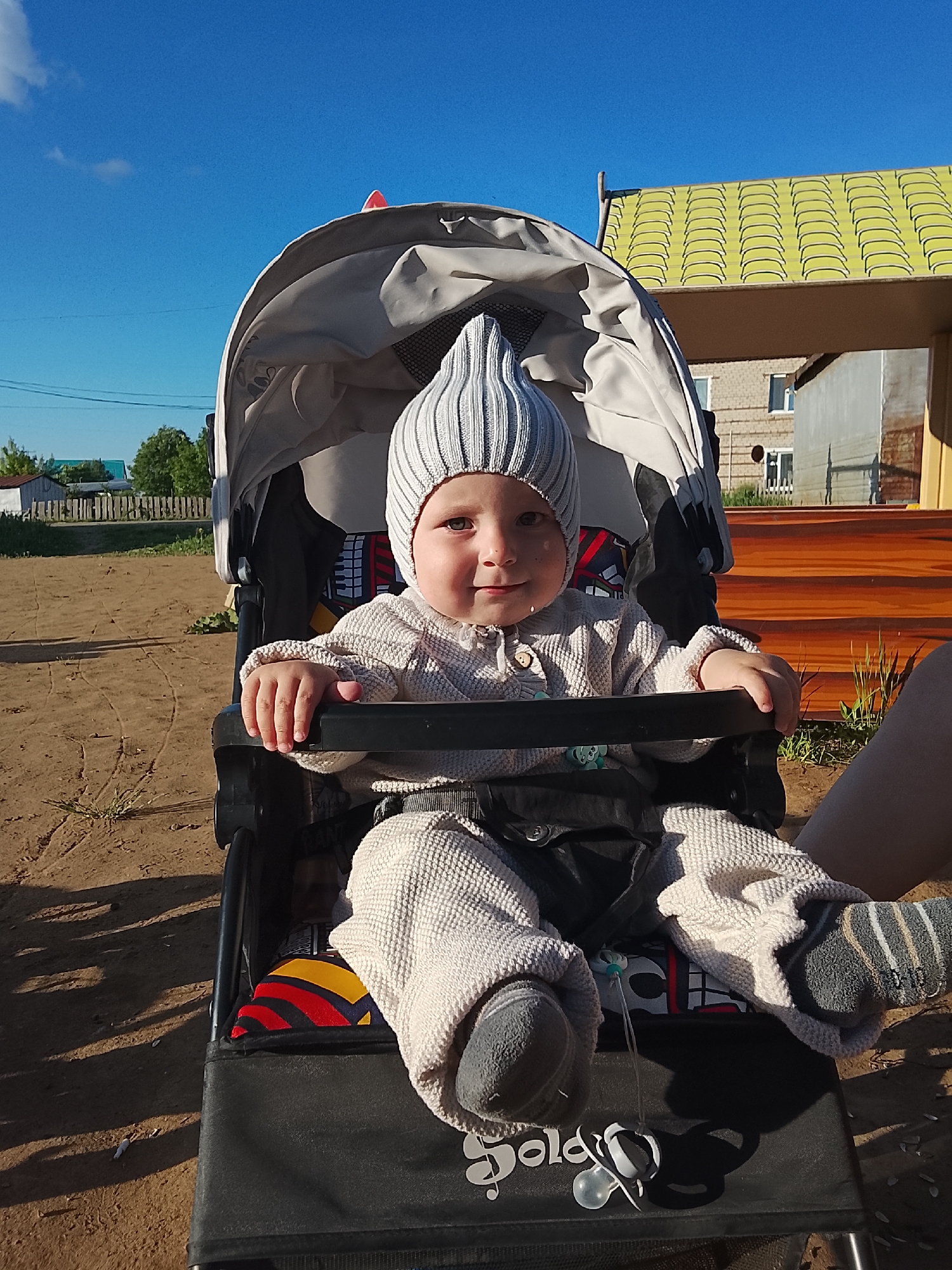 A baby sits in a stroller in Kuyeda, Russia. The baby wears a striped hat, a knitted sweater, and matching pants with socks. The stroller is black with a patterned seat and a grey canopy. A pacifier hangs from the stroller. Behind the stroller, there is a wooden structure with a green roof. The ground is dirt. The sky is clear and blue. In the background, there are houses and trees.