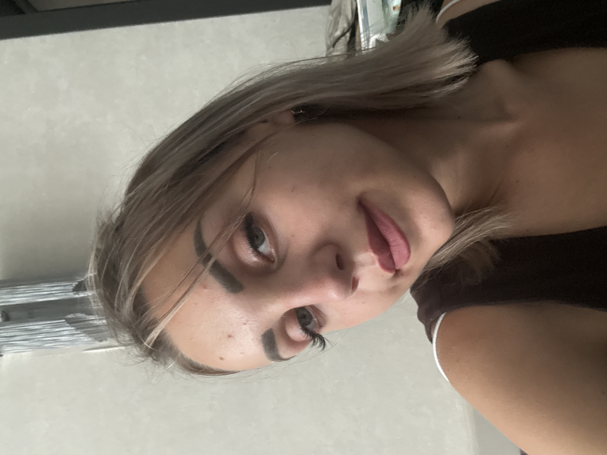 The image shows a close-up, waist-up portrait of a young woman with blonde hair and fair skin. She is wearing a dark brown tank top with white trim. She has makeup on, including eyeliner, mascara, and lipstick. Her hair is parted to her left and falls past her shoulders. The background is a light-colored wall with some vertical elements above her head, possibly a radiator or blinds. To the right of the woman, a dark object, potentially a piece of furniture or a doorway, is visible. There is no indication of the location being Yuzhno-Sakhalinsk, Russia.