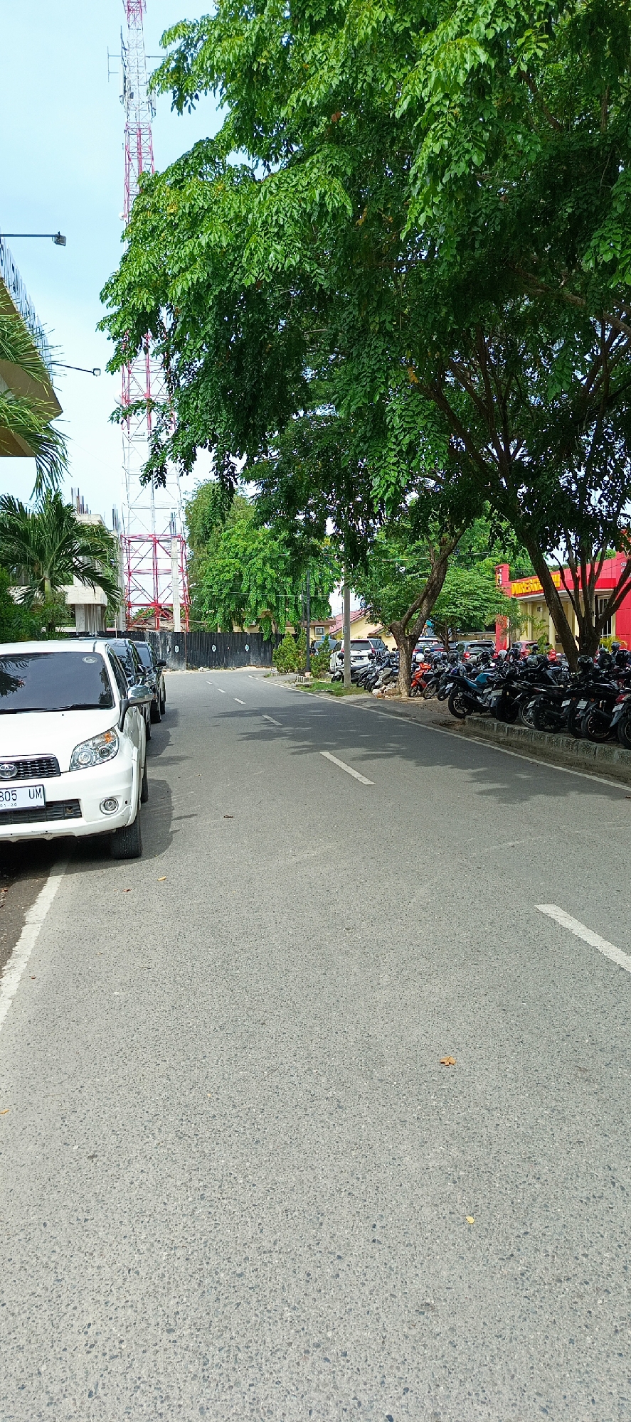 The image shows a street scene in Banda Aceh, Indonesia, likely in a commercial or public area. On the left side of the road, a white car is parked, followed by other vehicles. The road itself is paved and has white lane markings. To the right of the road, a large number of motorcycles are parked in a designated area. Beyond the motorcycles, there are buildings, including a red building with yellow accents. Trees with lush green foliage line the street, providing shade. In the background, a tall telecommunications tower is visible against a bright blue sky. The overall impression is a typical day in a busy urban environment in Banda Aceh.