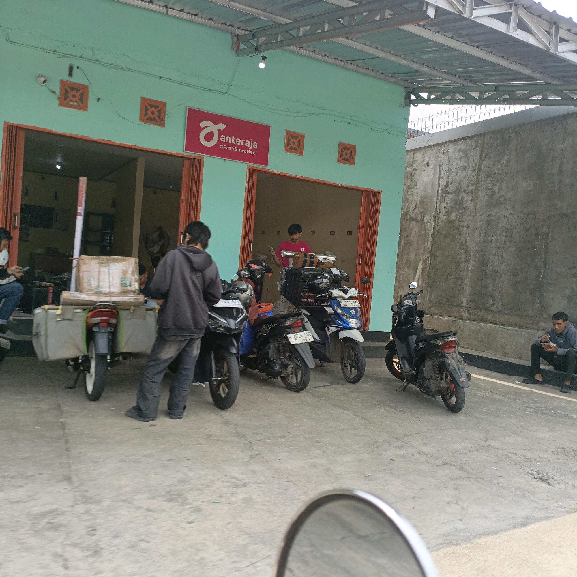 The image depicts an outdoor scene in front of a building with a light teal-colored facade and an open entrance. A red and white sign with the Anteraja logo and the hashtag #PastiBawaHepi is visible on the building. Several motorcycles are parked in front of the building, some with saddlebags and cargo.
In the foreground, a person wearing a dark gray hoodie and dark pants stands next to a motorcycle equipped with large side bags and a box of cargo. Another person, partially visible, is seated on a motorcycle on the left, looking at a phone. Inside the building entrance, a person in a pink shirt is visible, holding a phone. Further to the right, a person is seated on a low wall, eating from a container.
The environment appears to be a street or parking area with a concrete ground. A tall concrete wall runs along the right side of the image, and a metal roof structure extends over the building entrance. The overall scene suggests a delivery or logistics hub, likely belonging to the Anteraja company, a courier service in Indonesia. Given the presence of an Anteraja sign and the typical operations of such services, this location could be a drop-off or pick-up point within the city of Salatiga, Indonesia, where Anteraja operates.