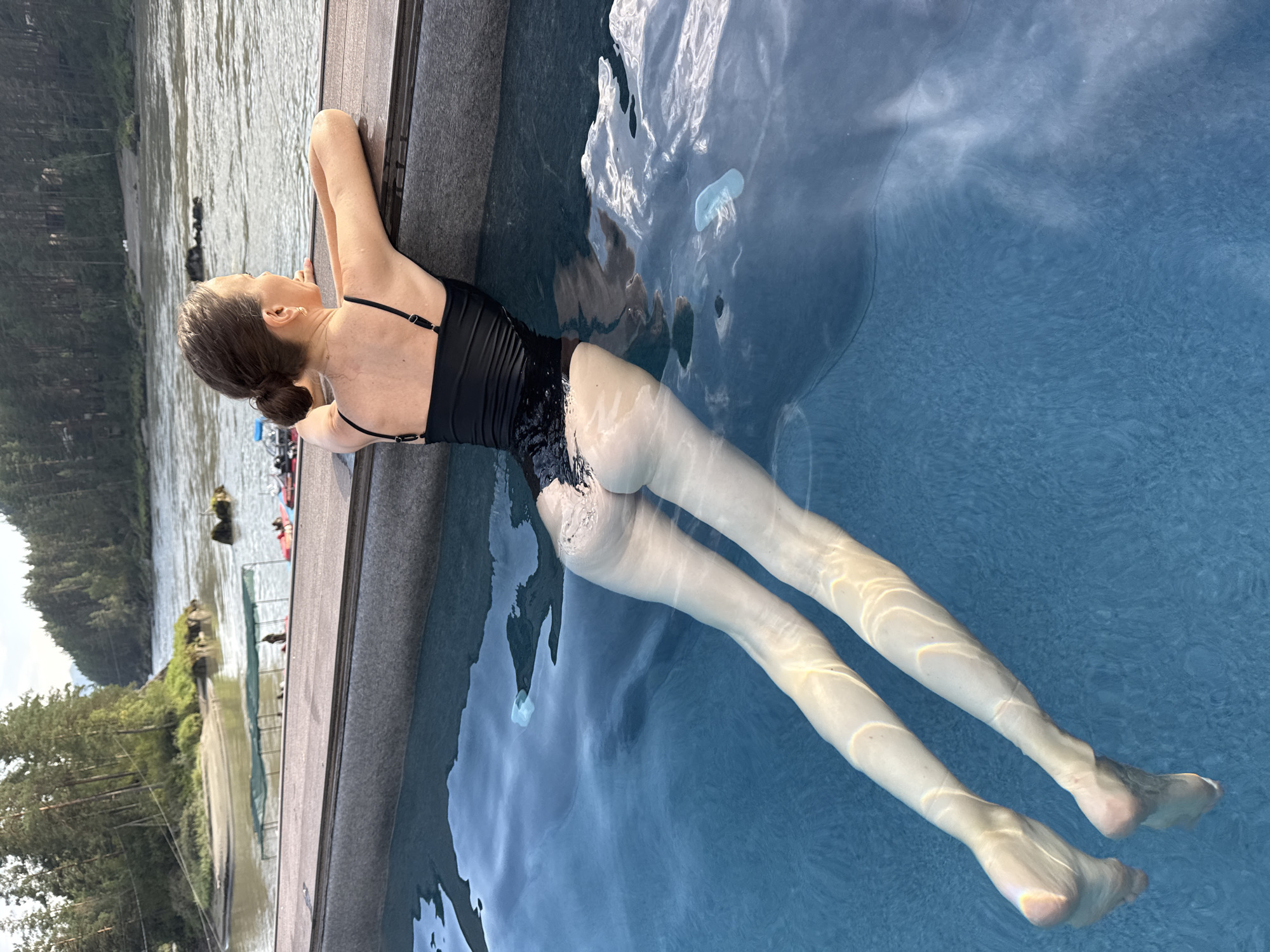 A woman in a black swimsuit leans on the edge of an infinity pool, her legs submerged in the water. The pool overlooks a wide, calm river surrounded by dense, green forest. The sky is overcast, suggesting either early morning or late evening. In the distance, a sandy bank can be seen, with a few small boats pulled ashore. The overall impression is one of serenity and natural beauty.