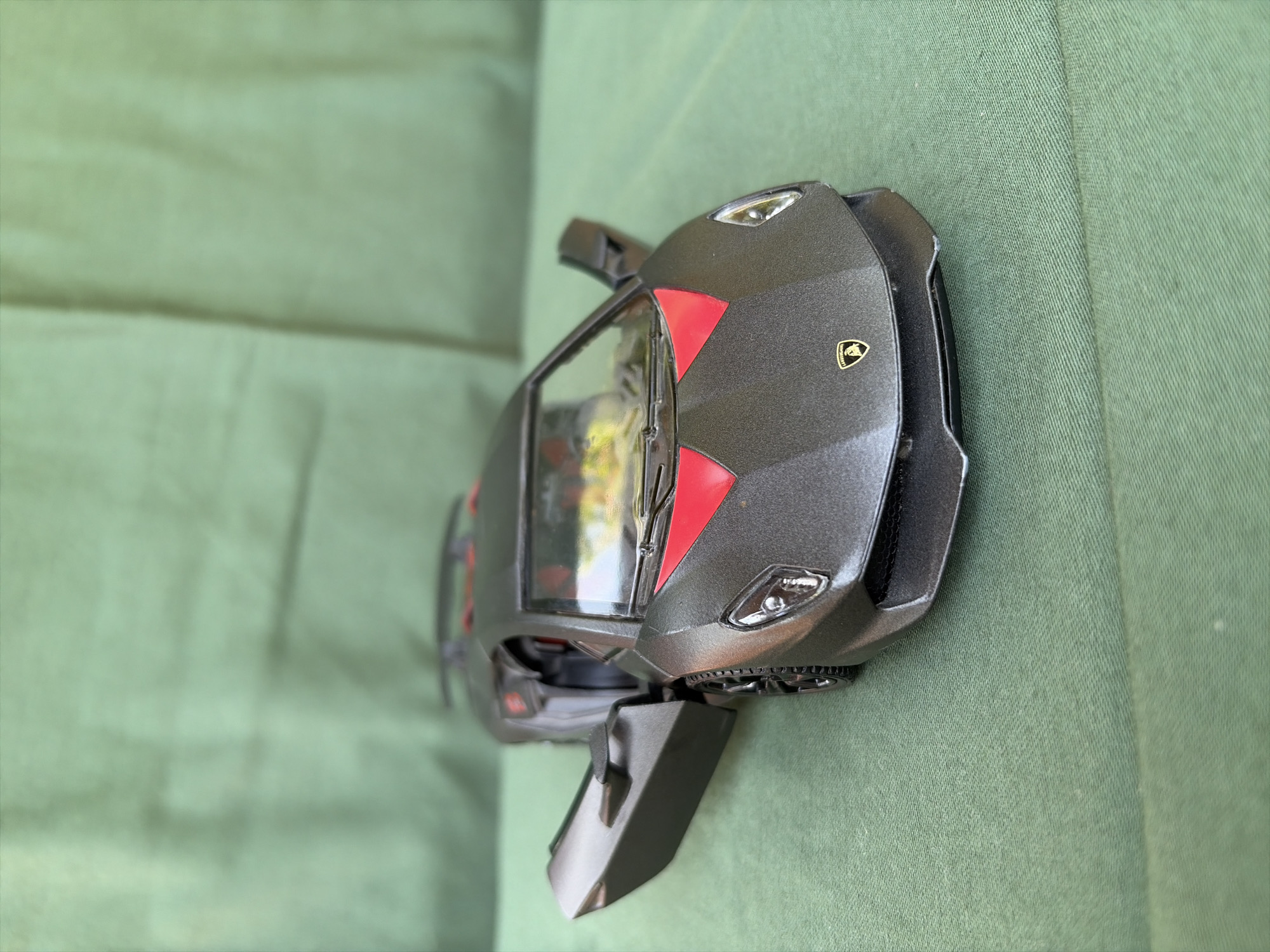A detailed, forensic, matter-of-fact description of the image, including the location in the city of Oxentea, Moldova:
The image shows a dark gray toy Lamborghini car with red accents on the hood and interior. The driver's side door is open, angled outward. The car is positioned on a textured green fabric surface, likely a tablecloth or backdrop. The background is a blurred green fabric, suggesting a draped setting. No people are visible in the image. The car has visible headlights, a windshield, wheels, and a small Lamborghini logo on the front. The overall lighting is natural, casting soft shadows. The environment appears to be indoors or in a sheltered outdoor space, with the green fabric serving as a consistent backdrop. The city of Oxentea, Moldova, is not depicted in this image.