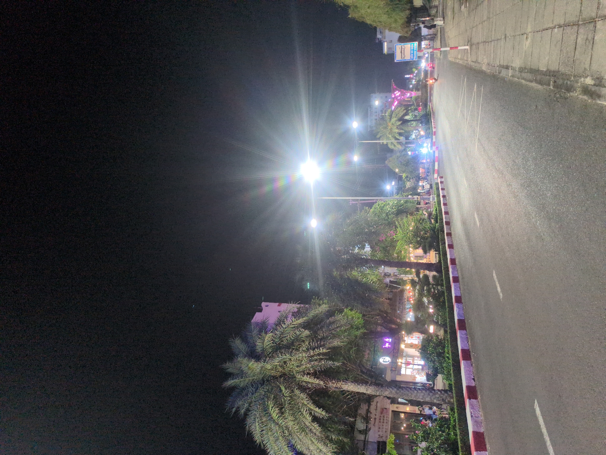 This is a nighttime view of a street in Tuy Hòa, Vietnam. The asphalt roadway runs vertically through the image, lit by bright streetlights. A raised median with trees and shops borders the road on the left side. There are people visible near the shops. Sidewalks line the road, with a concrete barrier between the road and sidewalk. A sign and pink structure can be seen further down the road. The sky is dark.