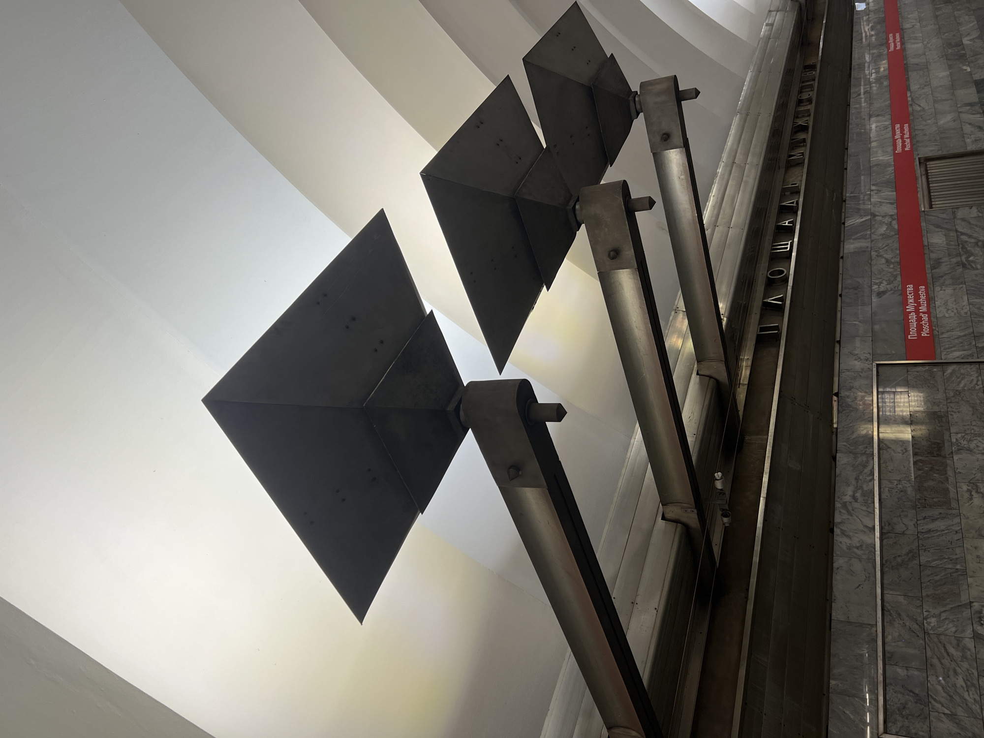 The image depicts the interior of a subway station in Saint Petersburg, Russia. The scene focuses on the ceiling architecture, which features a series of angular, dark-colored light fixtures suspended from metallic arms. These fixtures are positioned against a backdrop of a white, vaulted ceiling with curved lines. Below the ceiling, a metallic sign displays text in Cyrillic and Latin alphabets. The text reads "ПЛОЩАДЬ МУЖЕСТВА" and "Ploschad' Muzhestva". The lower portion of the image shows a tiled wall with a red stripe and more text. The lighting is ambient, casting shadows that highlight the geometric forms of the light fixtures and the curvature of the ceiling. The station appears to be empty of people.