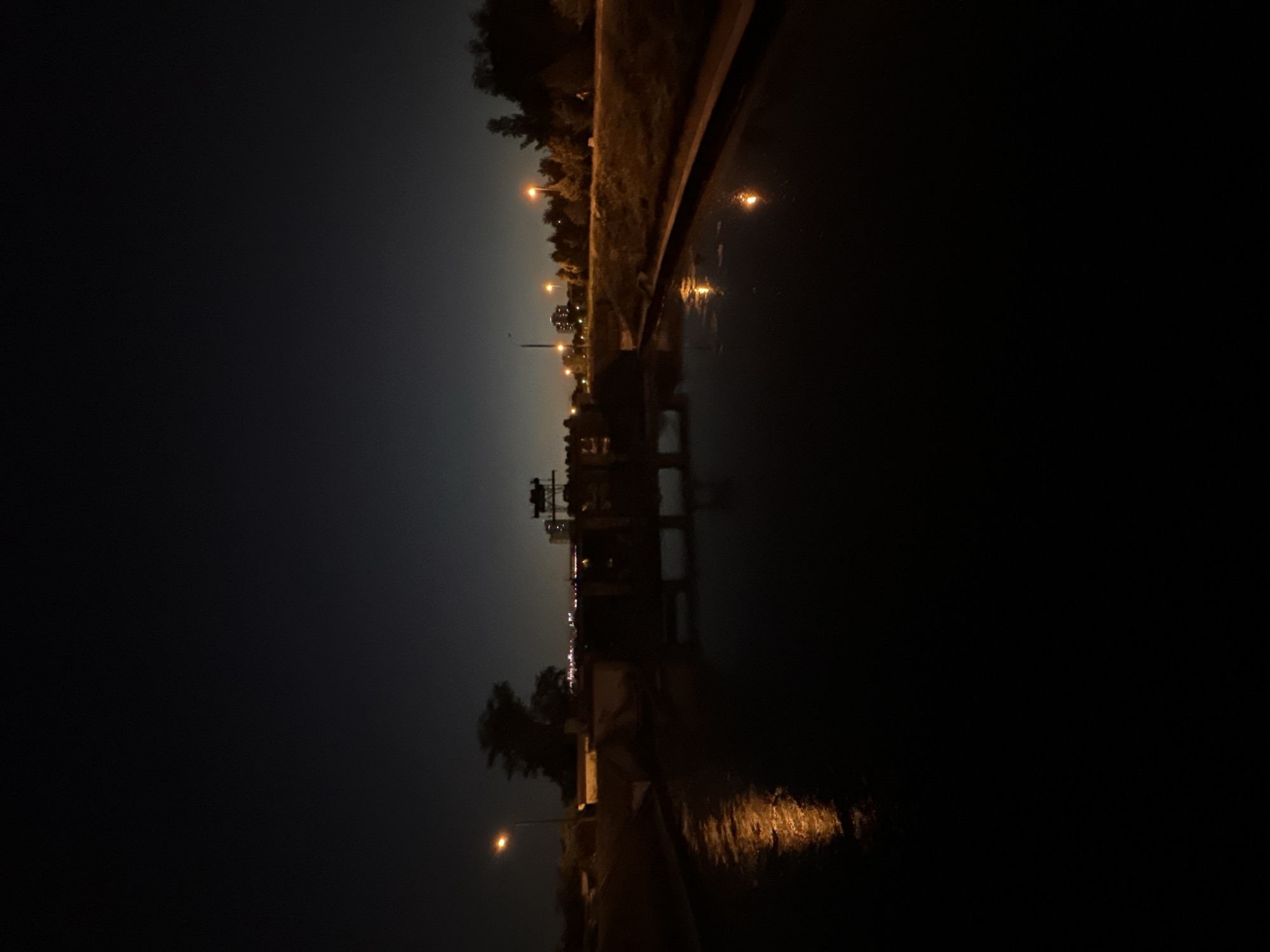 At night, a wide canal reflects the dark sky and distant city lights. On the left bank, a lamppost illuminates a wet embankment and a small building. Further along the canal, a lock gate structure is visible, with lights of various colors dotting the distant cityscape and trees lining the far bank. The water's surface is disturbed in places by what appears to be light reflections from streetlights. The scene is predominantly dark, with artificial lights providing the only illumination.
