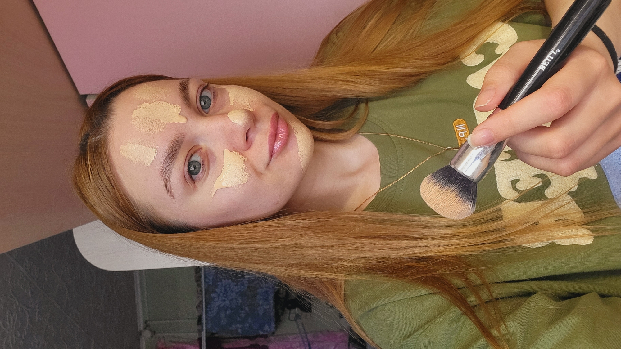 A young woman with fair skin and long, reddish-blonde hair is applying foundation to her face with a makeup brush. She has dabs of foundation on her forehead, cheeks, and nose. The brush is black with light brown bristles. The woman is wearing an olive green t-shirt and a gold necklace. The background consists of a pink surface and a glimpse of a room. The location is Зипуново, Russia.