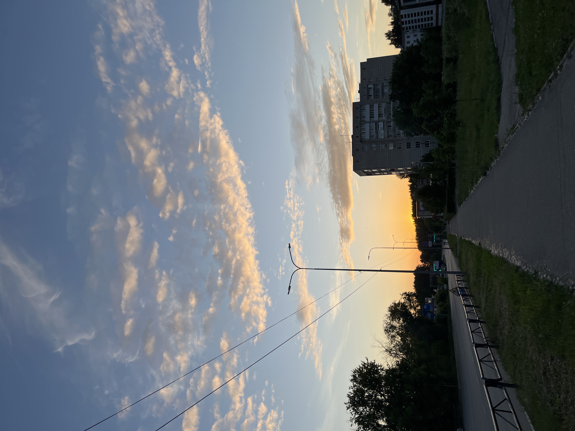 The image presents an urban street scene in Chaikovsky, Russia, captured at sunset. The sky transitions from a light blue at the zenith to a vibrant orange-yellow at the western horizon, where the sun is partially visible as a bright orange disc behind distant trees. Scattered stratocumulus clouds, illuminated from below with warm, golden hues, are present across the sky. Multiple dark power lines diagonally traverse the upper and mid-sections of the frame, and several small birds are visible as dark specks against the sky.
In the foreground and midground on the left, an asphalt road is bordered by a dark metal fence featuring triangular patterns, with green grass between the fence and the roadway. Dense green trees line the far side of the road, beyond which a blue sign and a larger billboard structure are partially discernible. A green traffic light is visible further down the road, and a single, indistinct human figure is present near a streetlight on the left side of the road. Several tall, dual-lamp streetlights illuminate the road.
On the right side of the image, a paved sidewalk runs parallel to the road, separated by a strip of green grass. Beyond the sidewalk, a large, multi-story (approximately 9-10 floors) grey concrete residential building, characteristic of post-Soviet architecture with numerous balconies and windows, occupies a significant portion of the right midground and background. Smaller trees and patches of green grass are situated between the sidewalk and the building. Additional, less distinct buildings and trees are visible in the far right background.