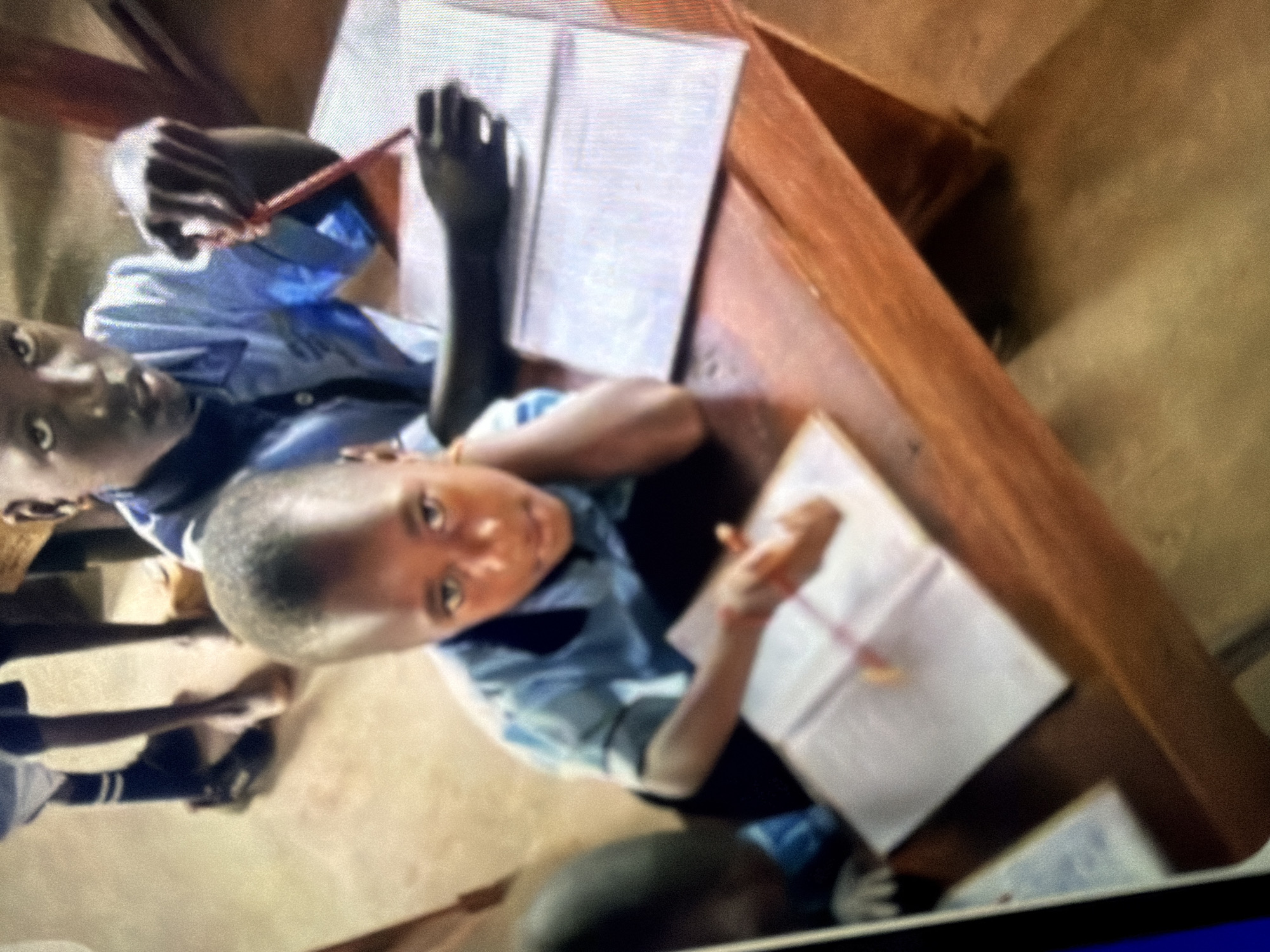 The image shows a close-up of two young boys sitting at a wooden desk, likely in a classroom. The boy in the foreground, facing the camera, has dark skin, short hair, and is wearing a light blue collared shirt. He is looking directly at the camera with a serious expression, resting his chin on his hand. In front of him is an open notebook with a pink pencil. To his left, another boy, also with dark skin and short hair, is wearing a similar light blue shirt. He is partially visible, focusing on an open notebook in front of him, with a red pencil in his right hand. The desk is made of polished wood, and the background appears to be a classroom setting with a light-colored floor. The lighting is subdued, suggesting an indoor environment.