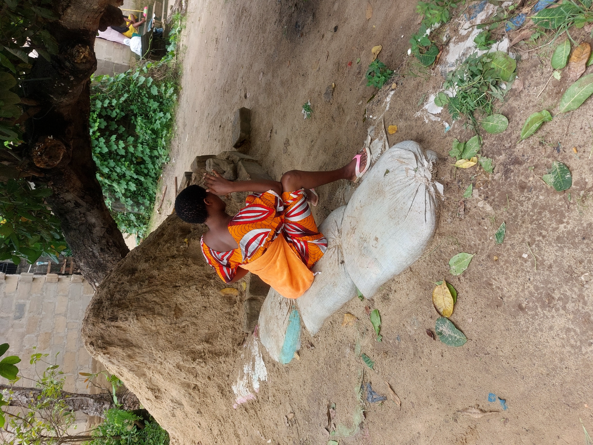 A young person wearing an orange and white patterned cloth sits on a pile of sandbags near a tree. The ground is sandy and has leaves scattered on it. The location appears to be outdoors in Dar es-Salaam, Tanzania, based on the vegetation and the style of dress.