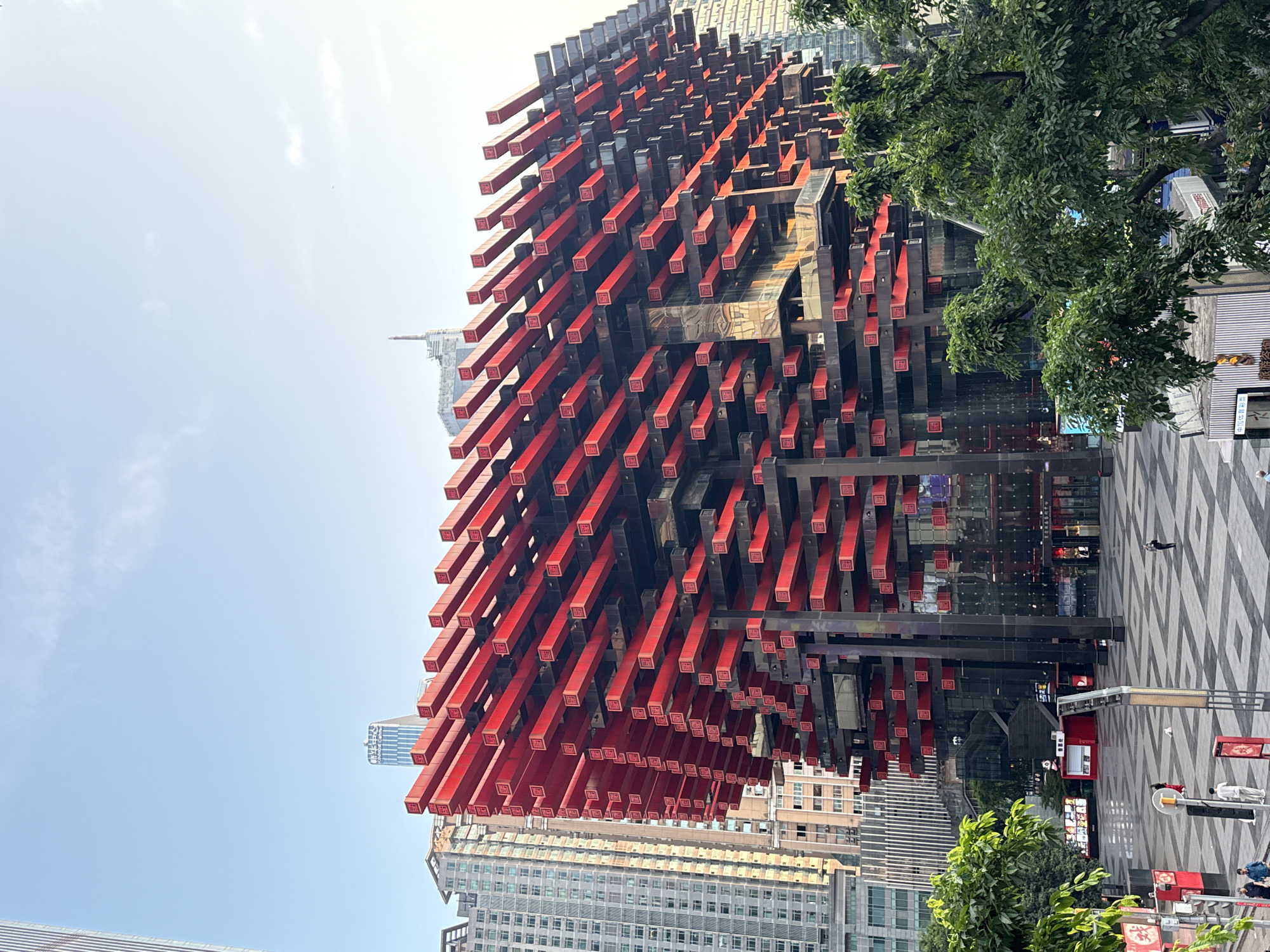 The image shows a low-angle view of the Raffles City Chongqing complex in Yuzhong District, China. The prominent feature is the horizontal red and black beams that make up part of the structure, which gives the impression of a building adorned with numerous red chopsticks or tubes. In the background, other high-rise buildings are visible, suggesting a dense urban environment. A small number of people are seen walking on a patterned grey and white pavement near the building. A tree with green foliage is visible on the right side of the frame. The sky is mostly clear.