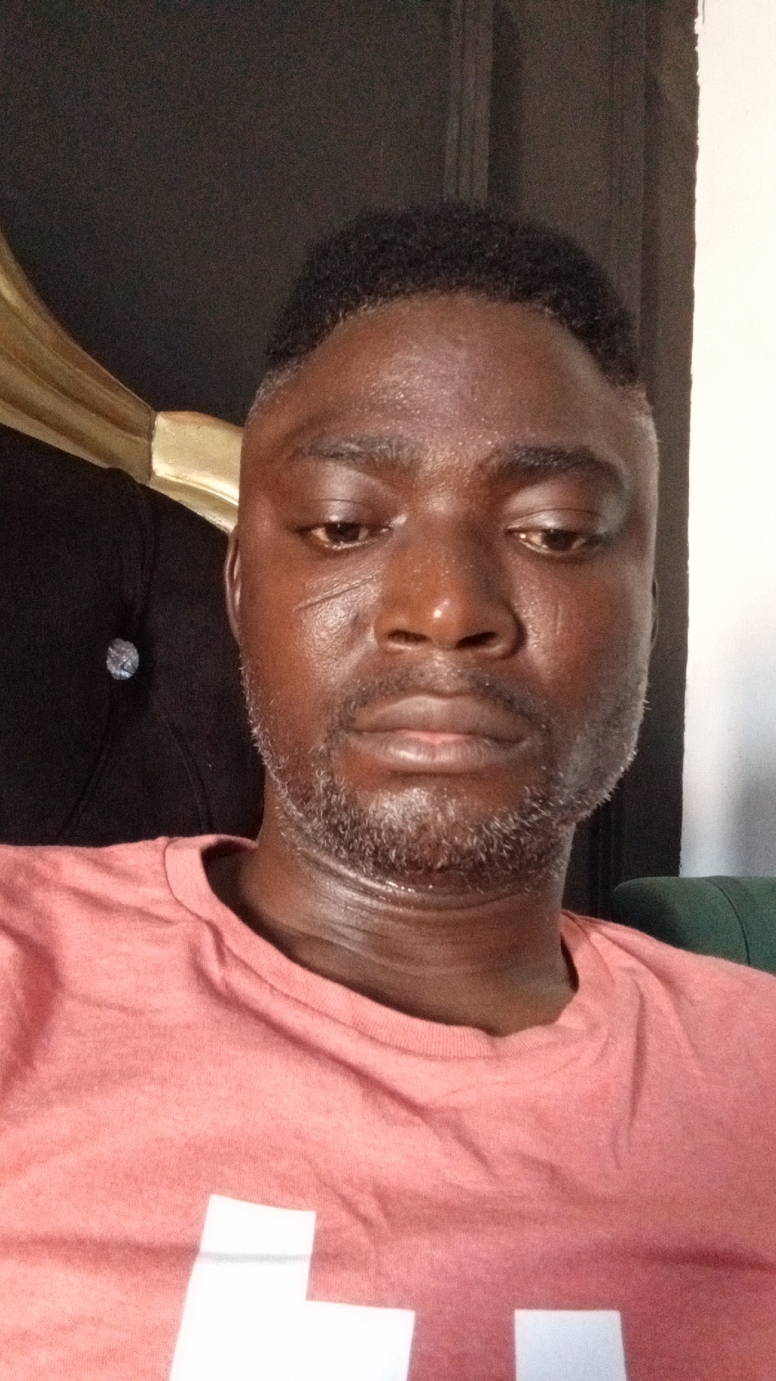 A close-up photograph shows a Black man wearing a salmon-colored t-shirt with a white design. The man has short, dark hair and a short, graying beard. He is looking downwards with a somber expression. A scar is visible on his left cheek. He is sitting on a black tufted chair with gold trim visible behind his head. The photo was likely taken indoors and, based on the request, is assumed to be in Sabon Tasha, Nigeria.