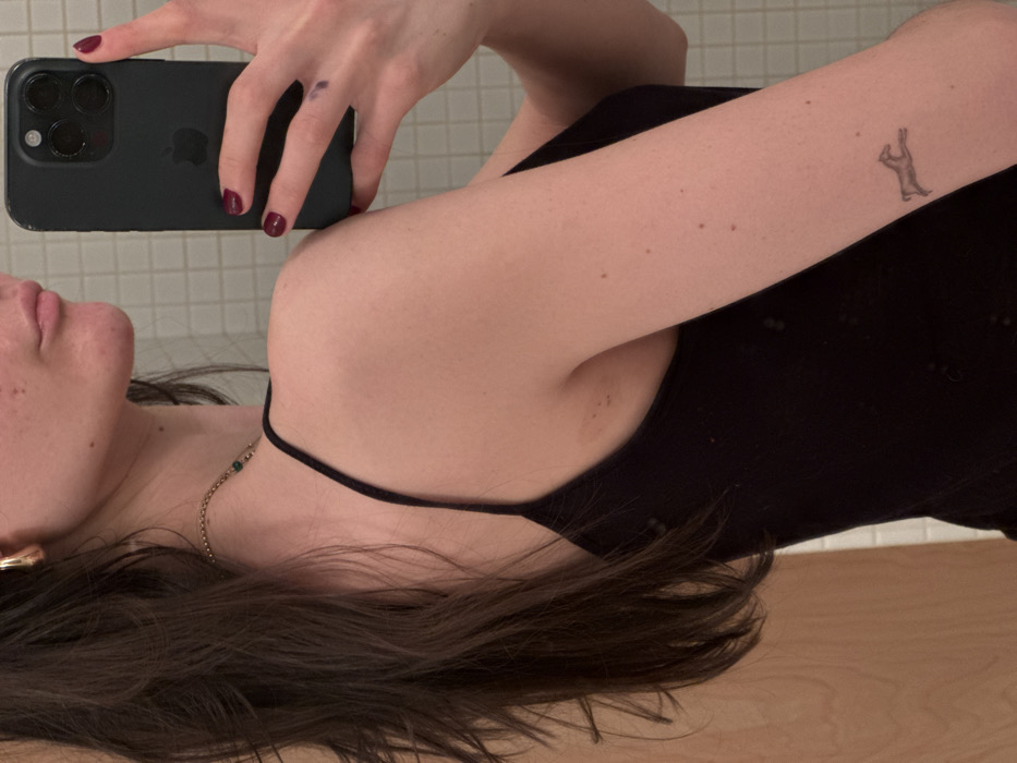 The image displays a mirror selfie of an individual, primarily showing their right profile from the upper torso upward. The subject possesses long, dark brown hair and fair skin. They are wearing a black spaghetti-strap garment, a gold-colored hoop earring in the right ear, and a delicate gold-colored chain necklace with a small green bead. A small, dark-colored tattoo depicting a galloping quadruped, resembling a deer or horse, is visible on the outer right forearm. The subject's right hand, adorned with deep red nail polish and a faint purple smudge near the index finger, holds a dark-colored smartphone, presumed to be an iPhone, positioned to capture the reflection. The background behind the subject consists of light-colored, possibly off-white or beige, square tiles forming a grid pattern, with a blurred vertical wooden surface visible on the far left. No discernible features are present to specifically identify a location within the city of New York, United States of America.