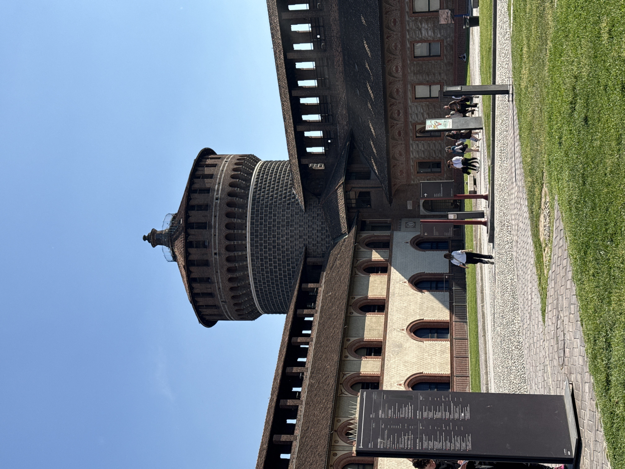 A sunny, outdoor wide shot captures a large, historic castle complex with several visitors. The image is dominated by a grand brick building featuring prominent towers and fortified walls. On the left, a tall, cylindrical tower with arched openings near its summit stands against a clear blue sky, characteristic of the Filarete Tower of Sforza Castle. To its right, a long section of the castle wall extends, showcasing intricate brick patterns and a row of smaller, rounded arched windows on a lower level, topped by battlements with rectangular openings.
In the foreground, a vibrant green grass lawn occupies the right side, while a paved walkway made of light-colored cobblestones runs along the base of the castle on the left. Several people are visible along this path. A small group stands in the middle ground, appearing to observe information boards or engage in conversation, while another individual walks further along the path. They seem to be tourists or visitors exploring the site.
A large, black information board stands prominently in the immediate foreground, covered with numerous lines of small white text that are unreadable. Another smaller, lighter-colored sign is visible further back, also unreadable. An arrow sign pointing left is visible on a pole near the path. The bright blue, cloudless sky and the strong shadows cast by the buildings and people indicate a clear, sunny day, likely late morning or early afternoon. The architectural style and setting strongly suggest the Sforza Castle (Castello Sforzesco) in Milan, Italy.