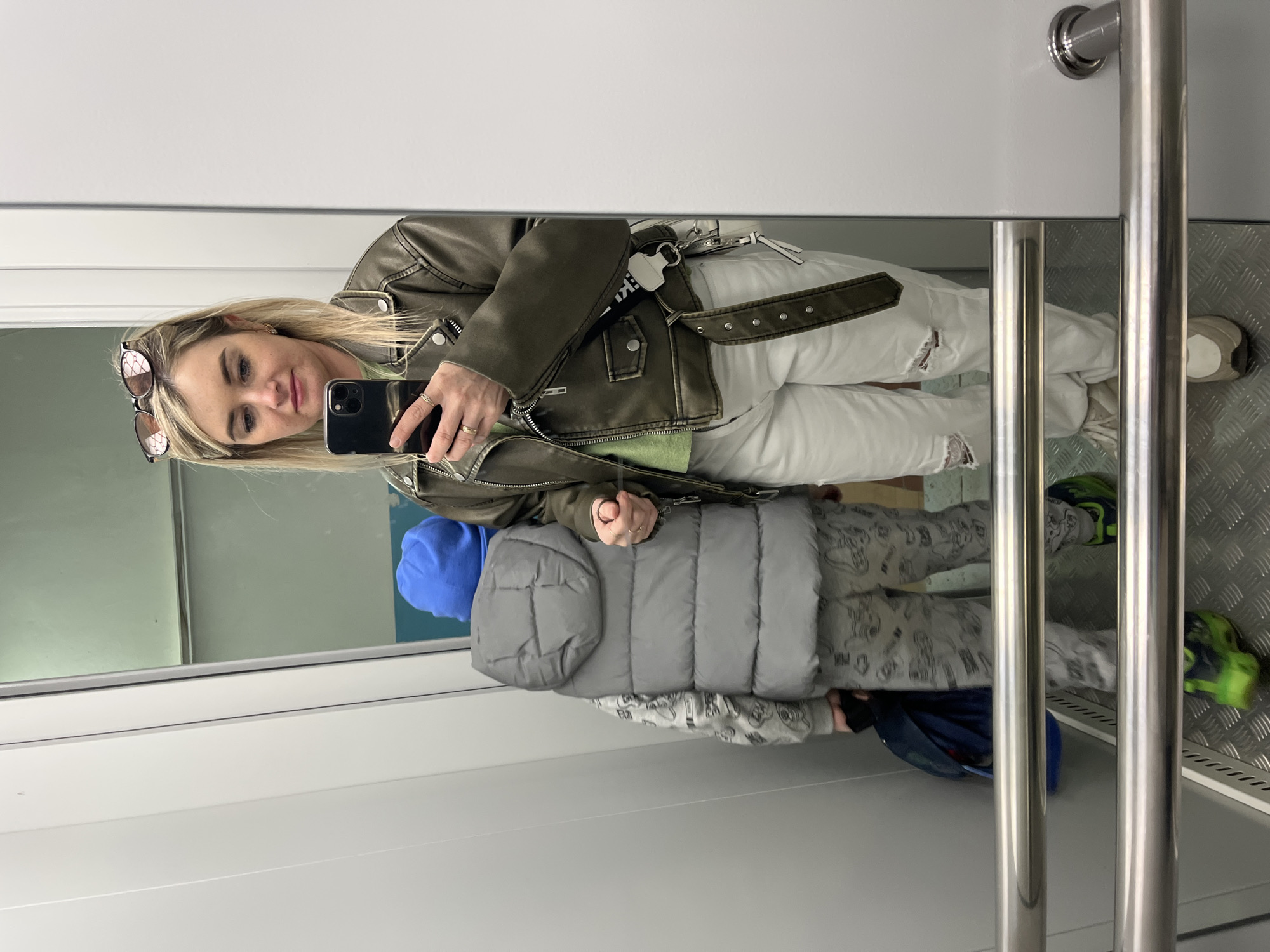 A woman and a child are reflected in a mirror. The woman is holding a phone and appears to be taking a selfie. She is wearing a light olive green leather jacket, white jeans with rips, and sunglasses perched on her head. The child is wearing a blue hooded sweatshirt and a grey puffer vest. They are standing inside what appears to be an elevator or a small enclosed space, as indicated by the metal walls and bars. The floor has a textured, metallic surface. The location cannot be determined based on the image alone. There is no mention of Russia or the city of Chaikovsky.