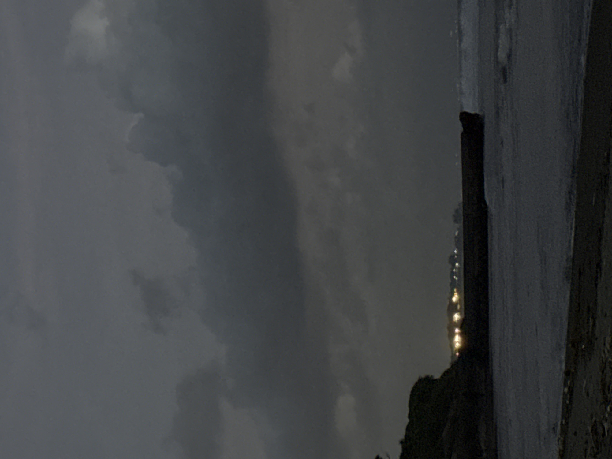 A nighttime view shows a beach and ocean under an overcast sky in Unknown, Unknown. The beach is in the lower right corner and stretches slightly towards the left. The ocean fills the right side of the image and appears dark and calm. A breakwater made of dark posts stands in the water. Lights flicker in the distance, possibly from a city. The sky is filled with heavy, grey clouds. Vegetation is visible on land.