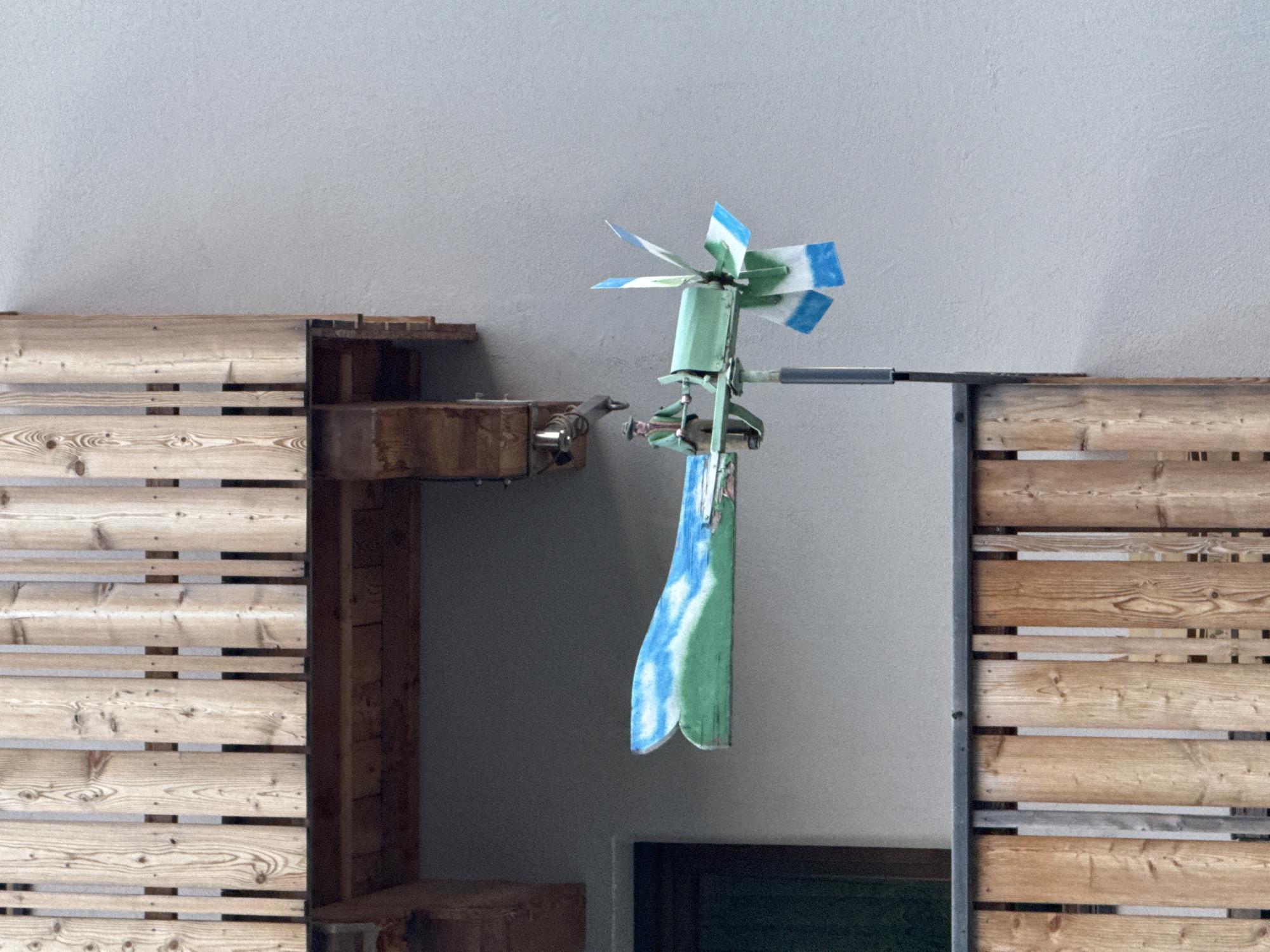 A miniature windmill decoration is mounted on a pole outside a building, likely a residential property, in La Ila - Stern - La Villa, Italy. The windmill is designed to resemble a person piloting a stylized airplane. The "plane" element is a long, narrow piece painted to depict a green landscape with a blue sky and white clouds. A small figure, dressed in a hat and green clothing, stands at the front of the "plane" as if steering it. The windmill's rotating blades are painted with alternating green and blue and white sections. The entire structure is positioned against a light gray textured wall, with a wooden balcony railing in the foreground and another wooden structure above, suggesting it's on a balcony or exterior wall of a multi-story building. A dark green door is visible in the background to the left.