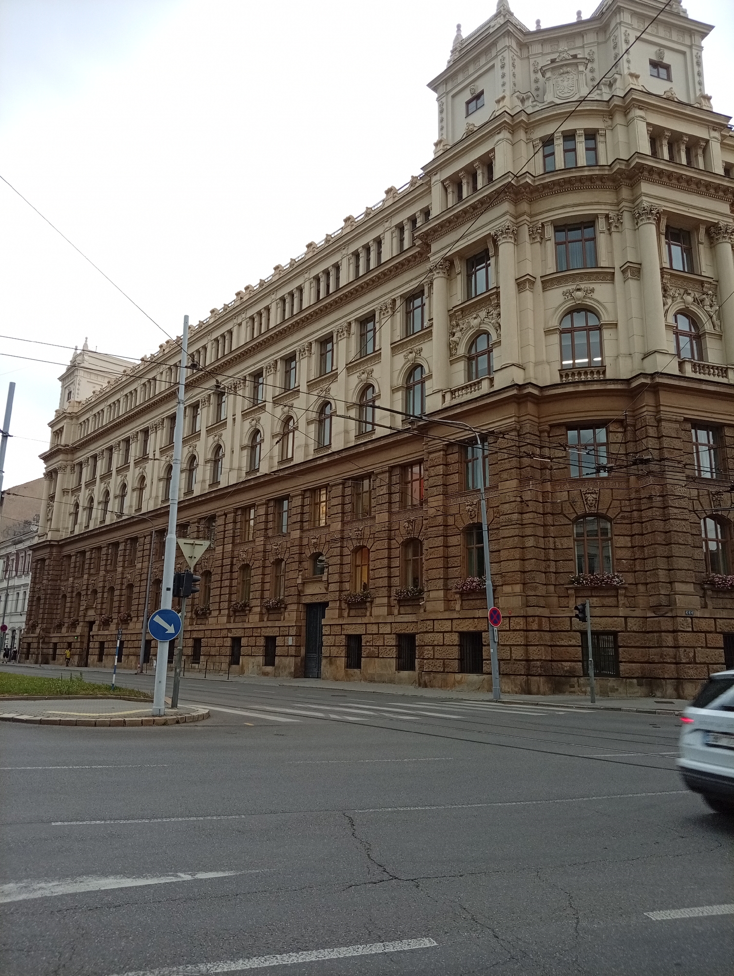 The image captures a street view in Brno, Czechia, focusing on the imposing facade of a grand building. The architecture is detailed, with a lighter-colored upper section featuring multiple rows of windows, ornate cornices, and classical columns. The lower section of the building is constructed from large, rough-hewn brown stones, adding a contrasting texture. Overhead, a network of utility wires crisscrosses the sky. In the foreground, a paved street is visible with road markings and a tram track. A blue directional sign indicating a right turn is mounted on a pole, alongside traffic lights and a red circular "no entry" sign. A white car is partially visible in the right side of the frame, in motion, and appears to be turning. The overall atmosphere is overcast, suggesting a cloudy day.